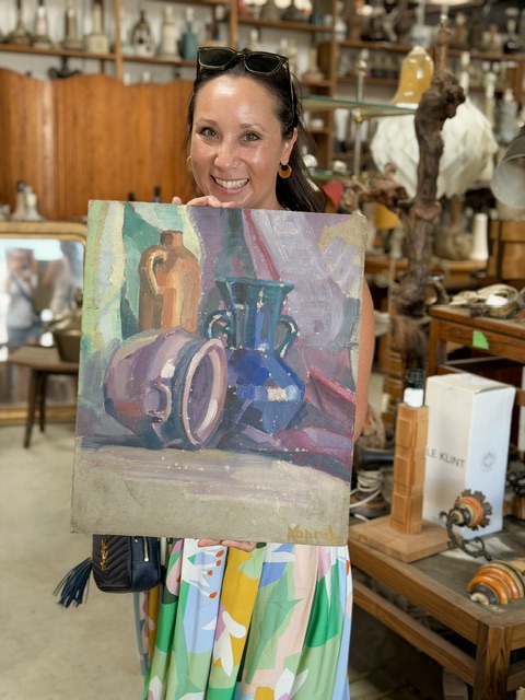 Woman holding a colorful still life painting of vases and bottles in an antique shop.