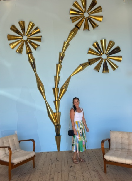 Woman standing in front of a wall with three large, gold, flower-shaped wall decorations, flanked by two chairs.