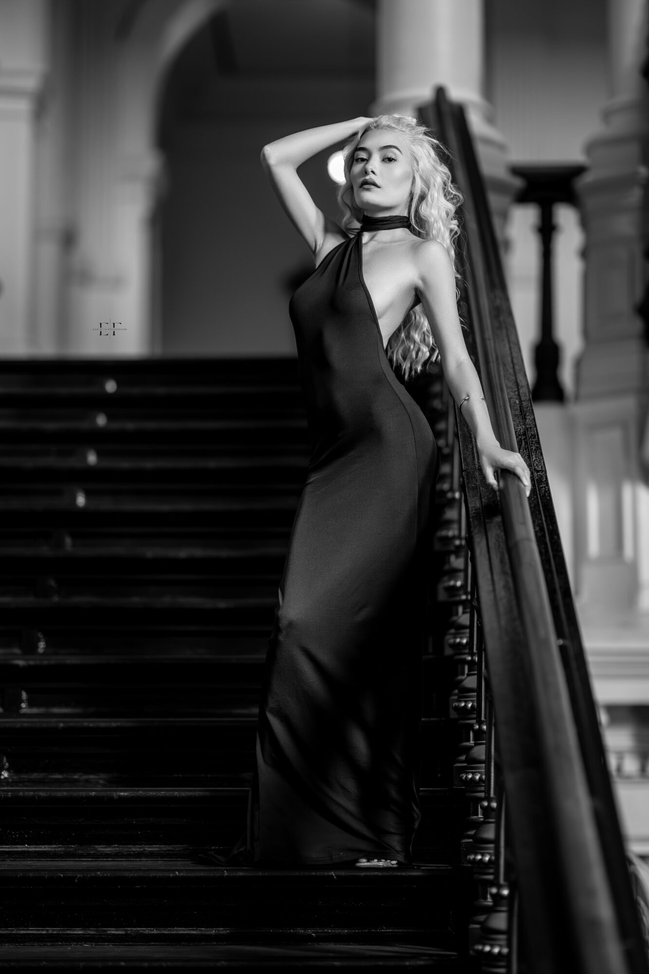 Woman in a long dress standing on staircase with hand on her head, elegant interior background, black and white.