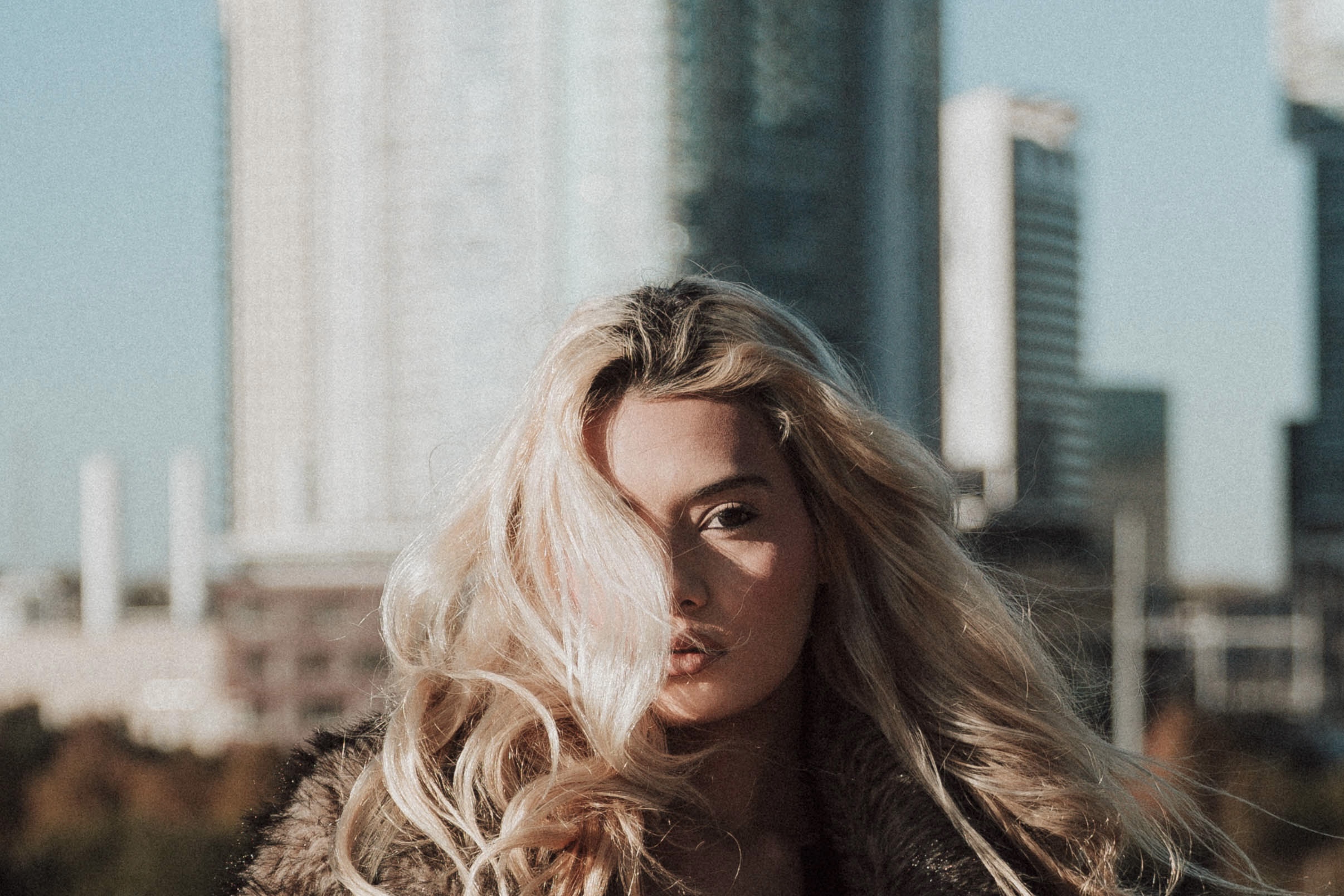 A woman with long, wavy blonde hair stands outdoors with a city skyline in the background.