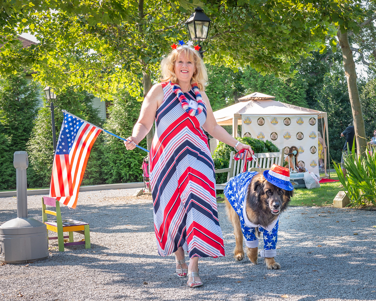 Woman walking a dog outdoors, holding an American flag, wearing patriotic clothing and a hat, with trees and a park setting.