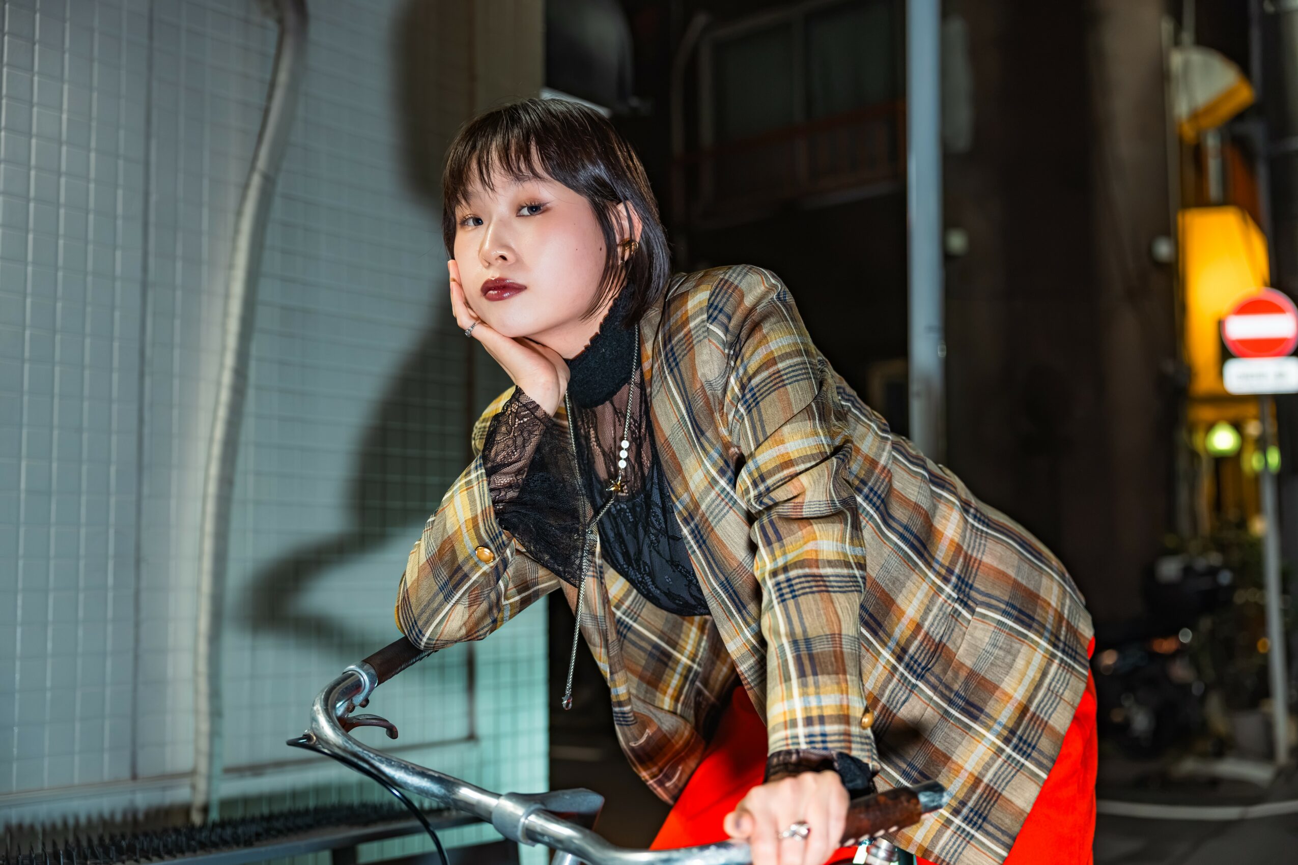 Young woman with short dark hair and makeup, wearing a plaid jacket, leaning on a bicycle handlebar at night.