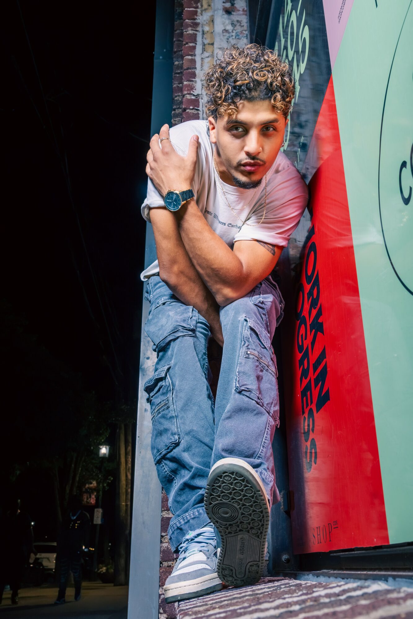 Young man with curly hair leaning against a colorful wall, looking at the camera, wearing a white t-shirt and jeans.