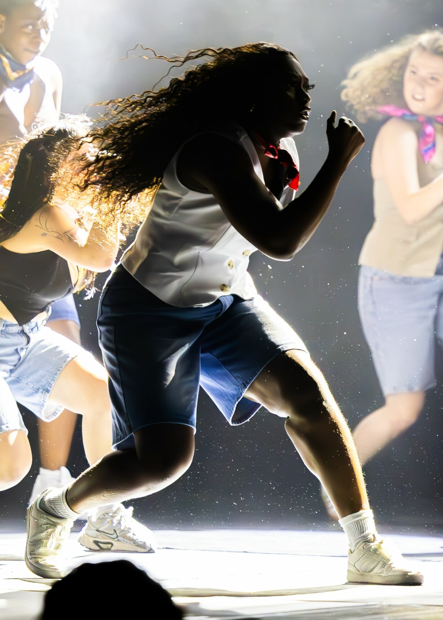 Person with long hair running or dancing, surrounded by other people, with dust or particles in the air, backlit scene.