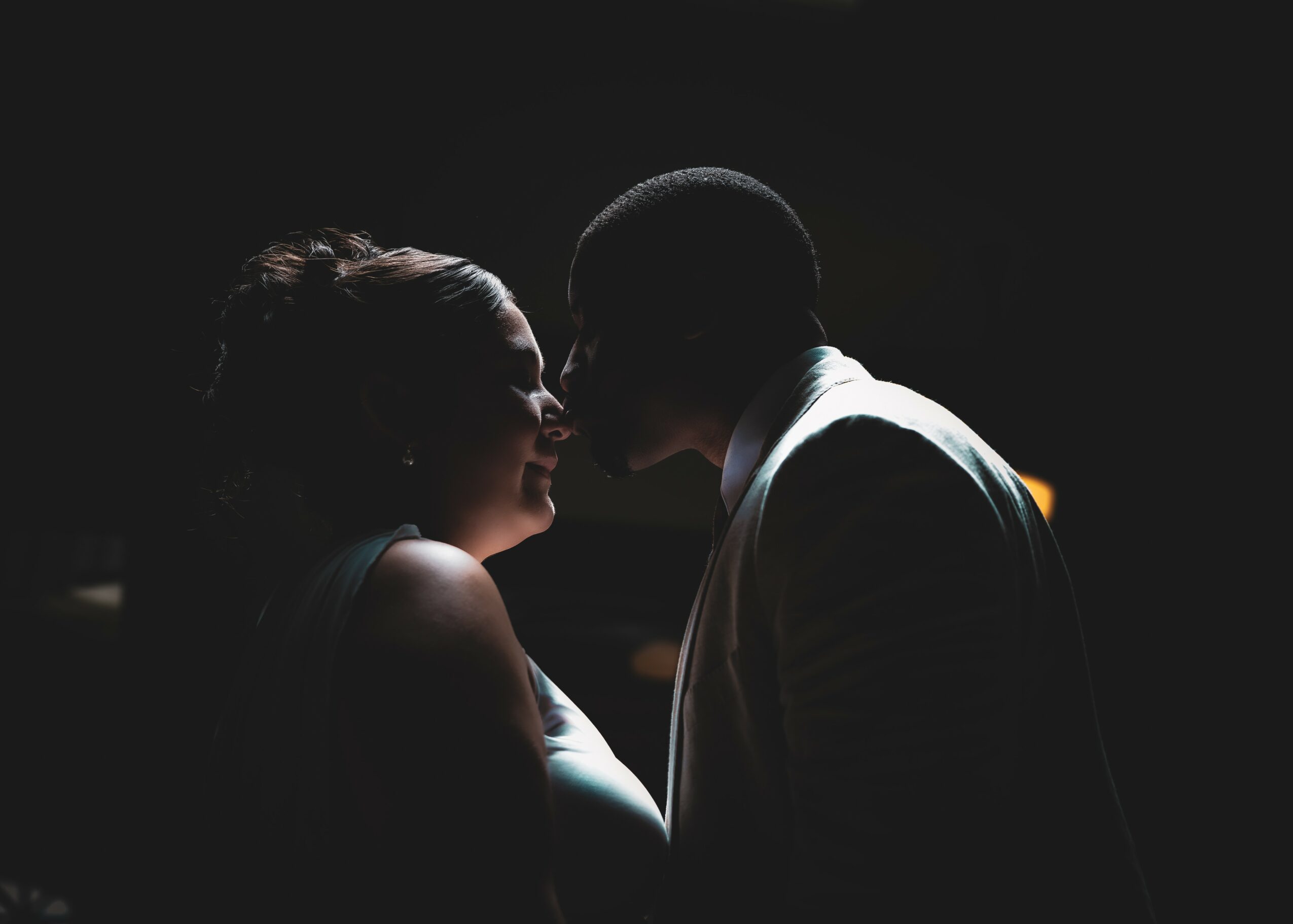Two people face each other closely in a dark setting, with their faces nearly touching, backlit silhouette.