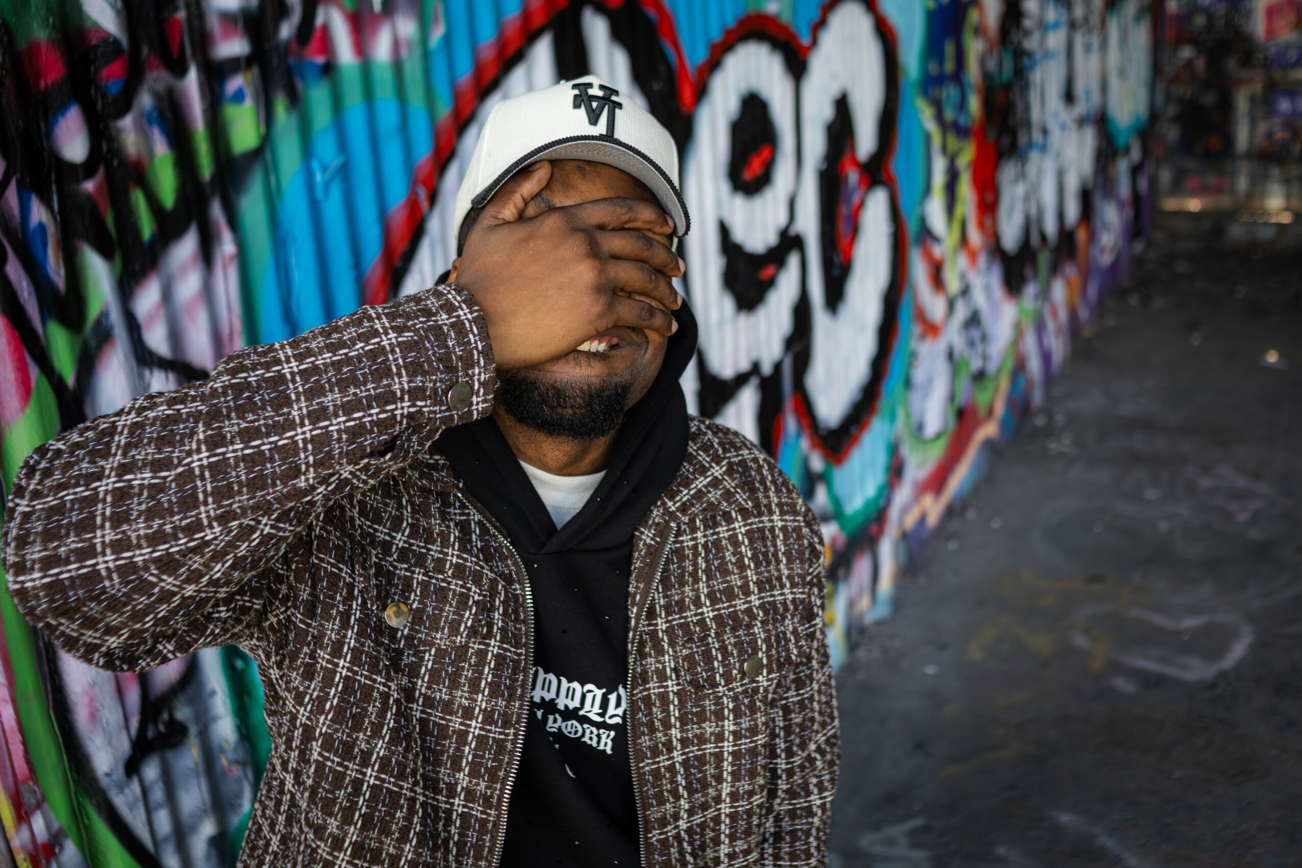 Person covering face with hand, standing in front of colorful graffiti wall, wearing a beanie and patterned jacket.