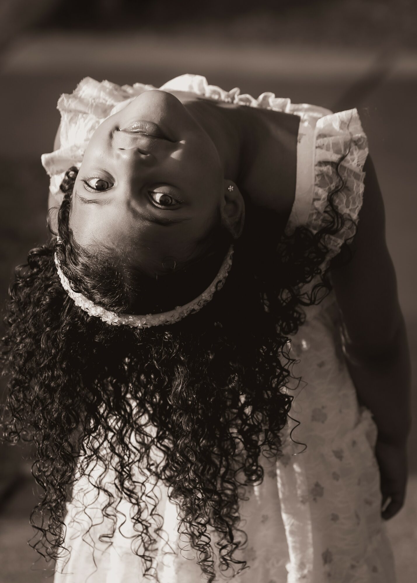 Young girl with curly hair and a headband, lying on her back, smiling, wearing a dress with ruffles.