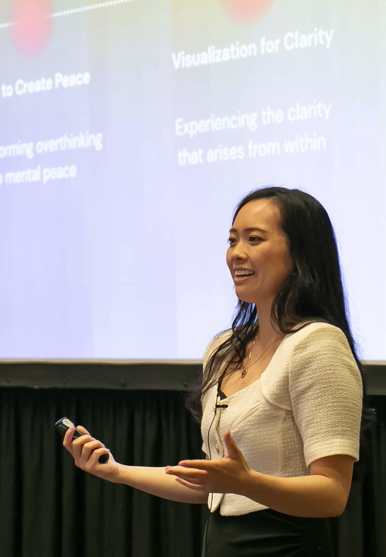 Woman smiling, holding a remote, standing in front of a presentation screen with text, gesturing with her left hand.