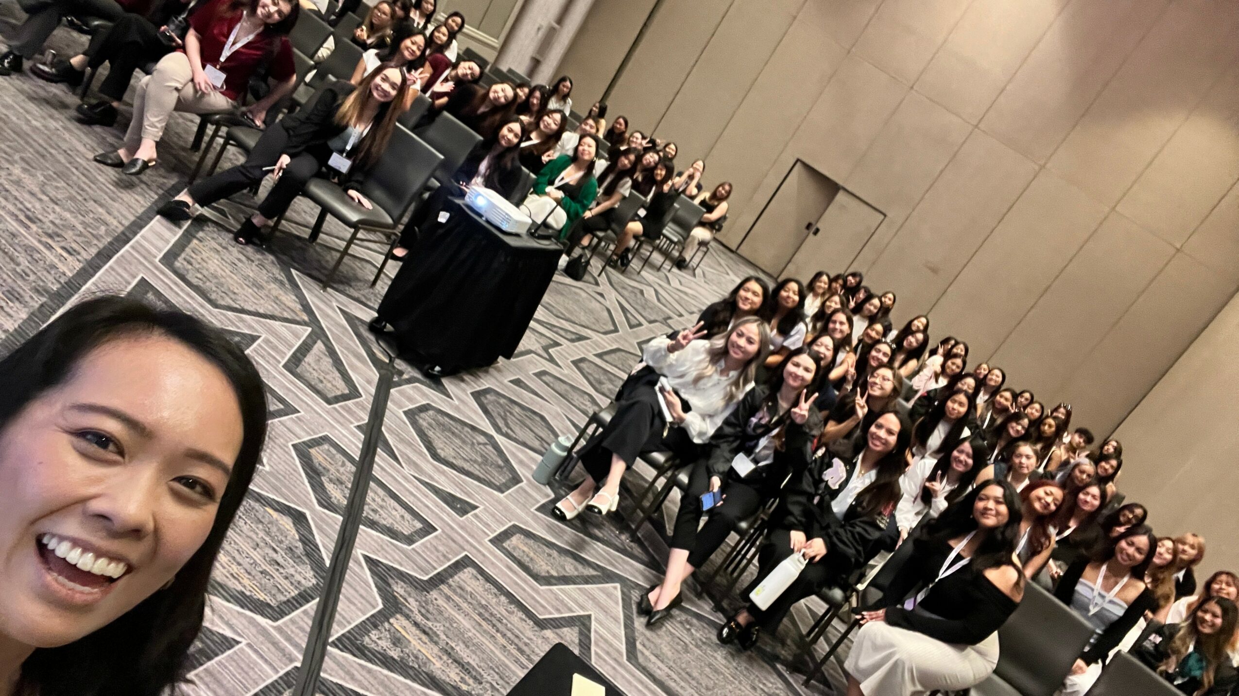 Group of people posing for a photo in a large room, with one person taking a selfie in the foreground.