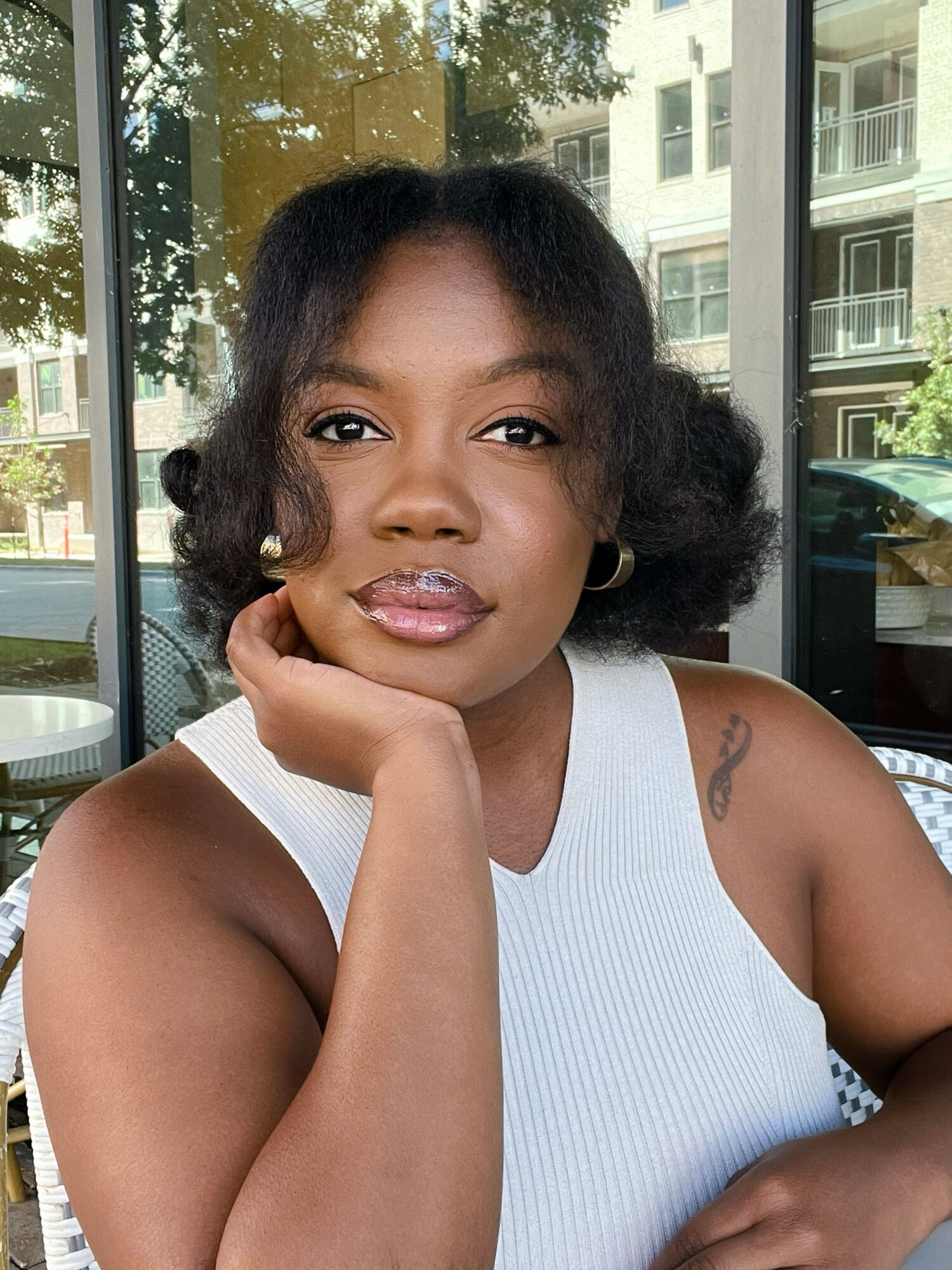 Woman with short curly hair wearing a white sleeveless top, resting her chin on her hand, sitting indoors near glass windows.