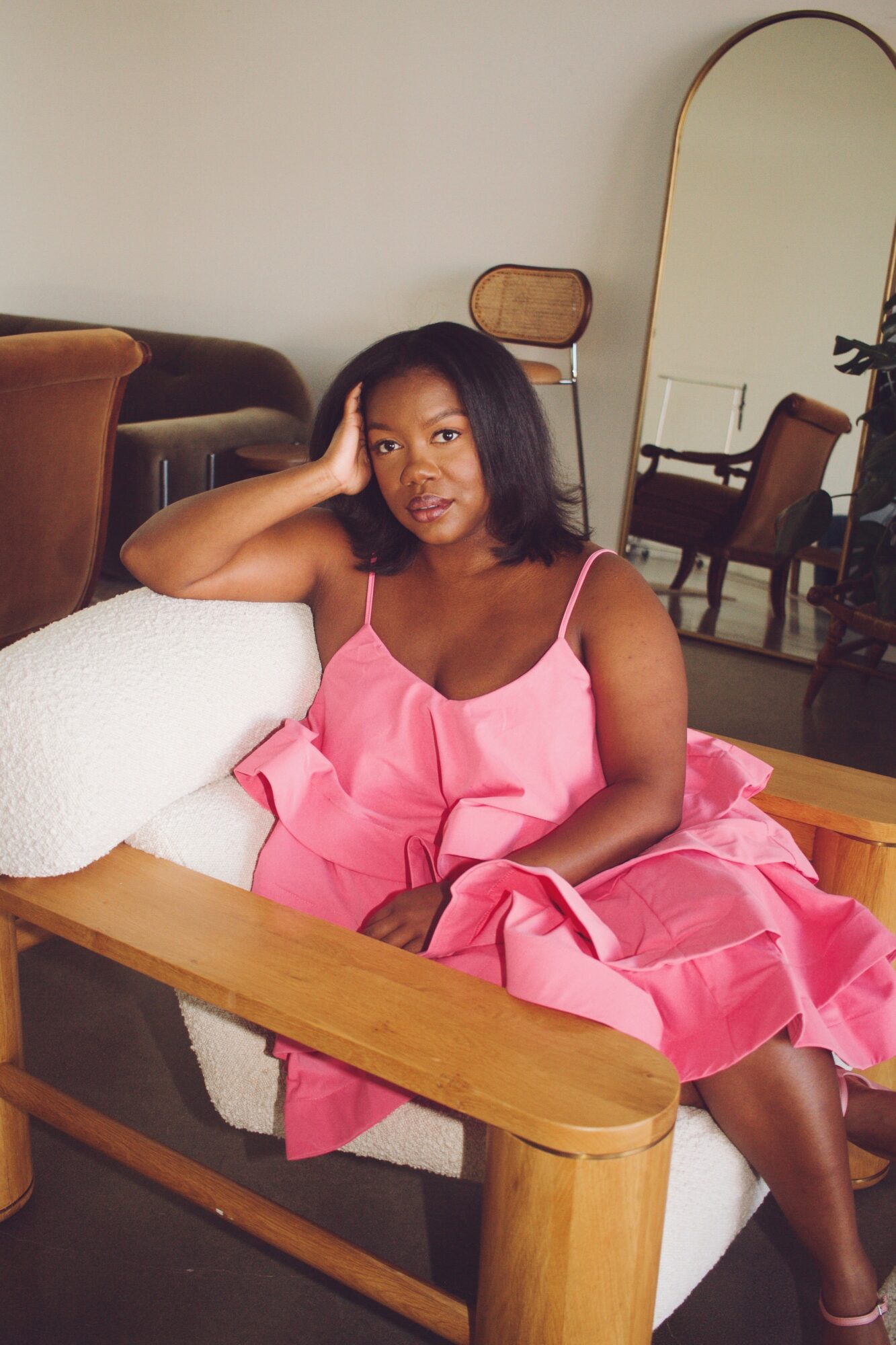 Woman in pink dress sitting on a wooden bench, resting her head on her hand, in a room with chairs and a mirror