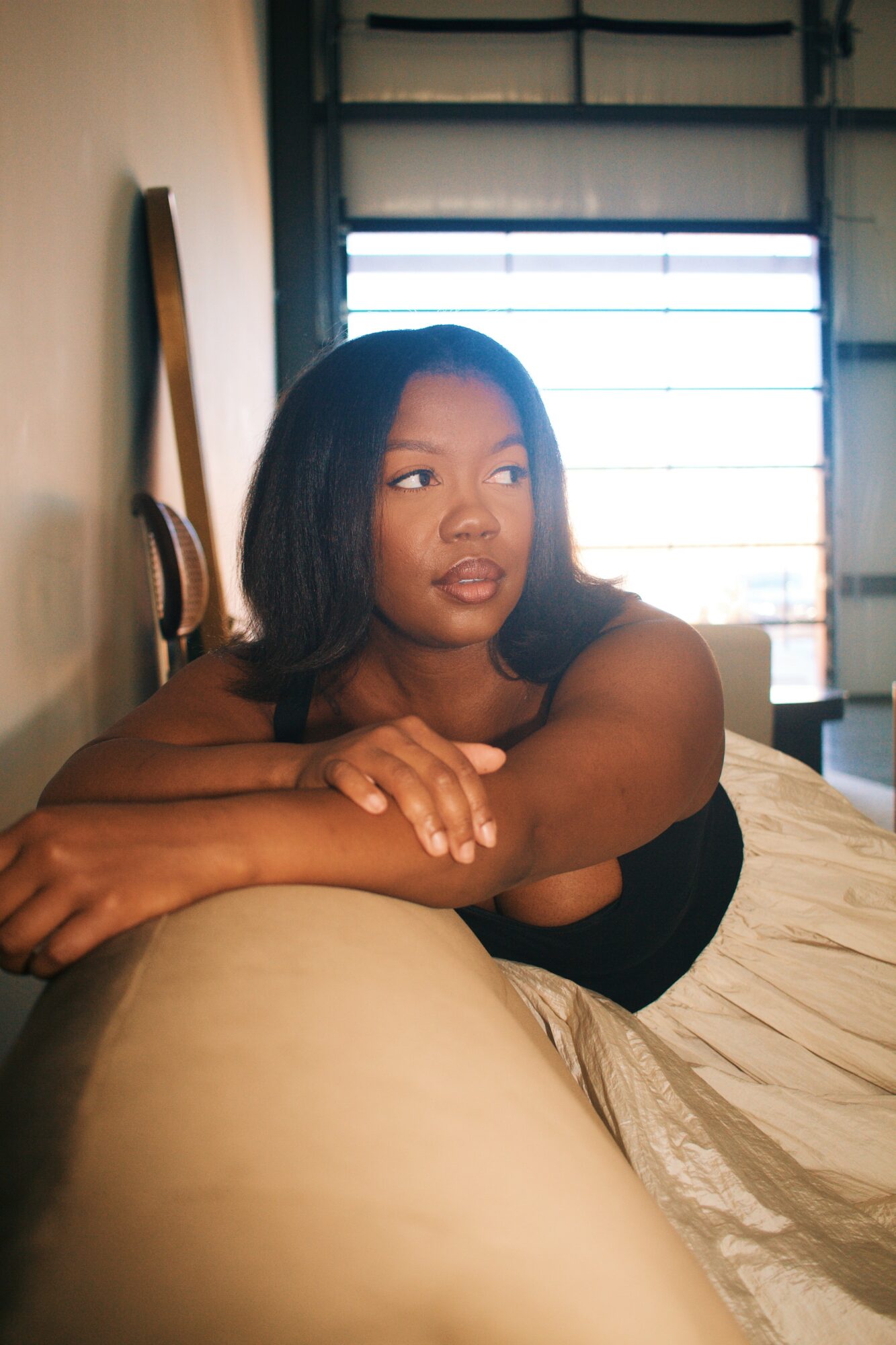 Young woman with shoulder-length hair resting on a bed, looking to the side near a window with blinds.