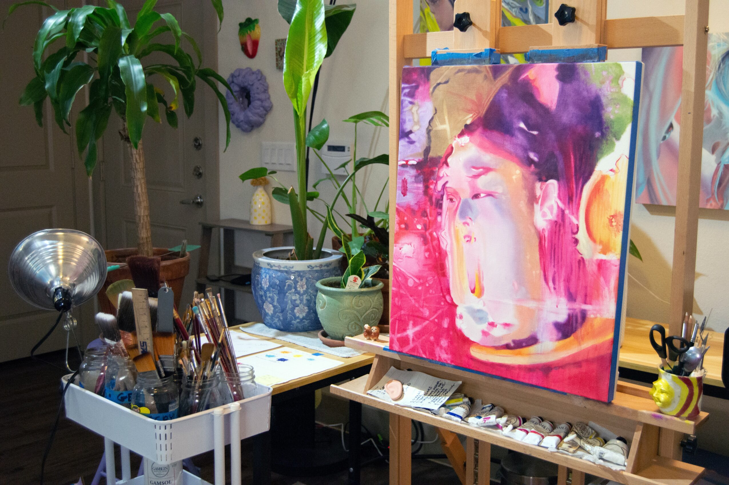 Art studio with a colorful abstract painting on an easel, plants, and art supplies on tables and shelves.