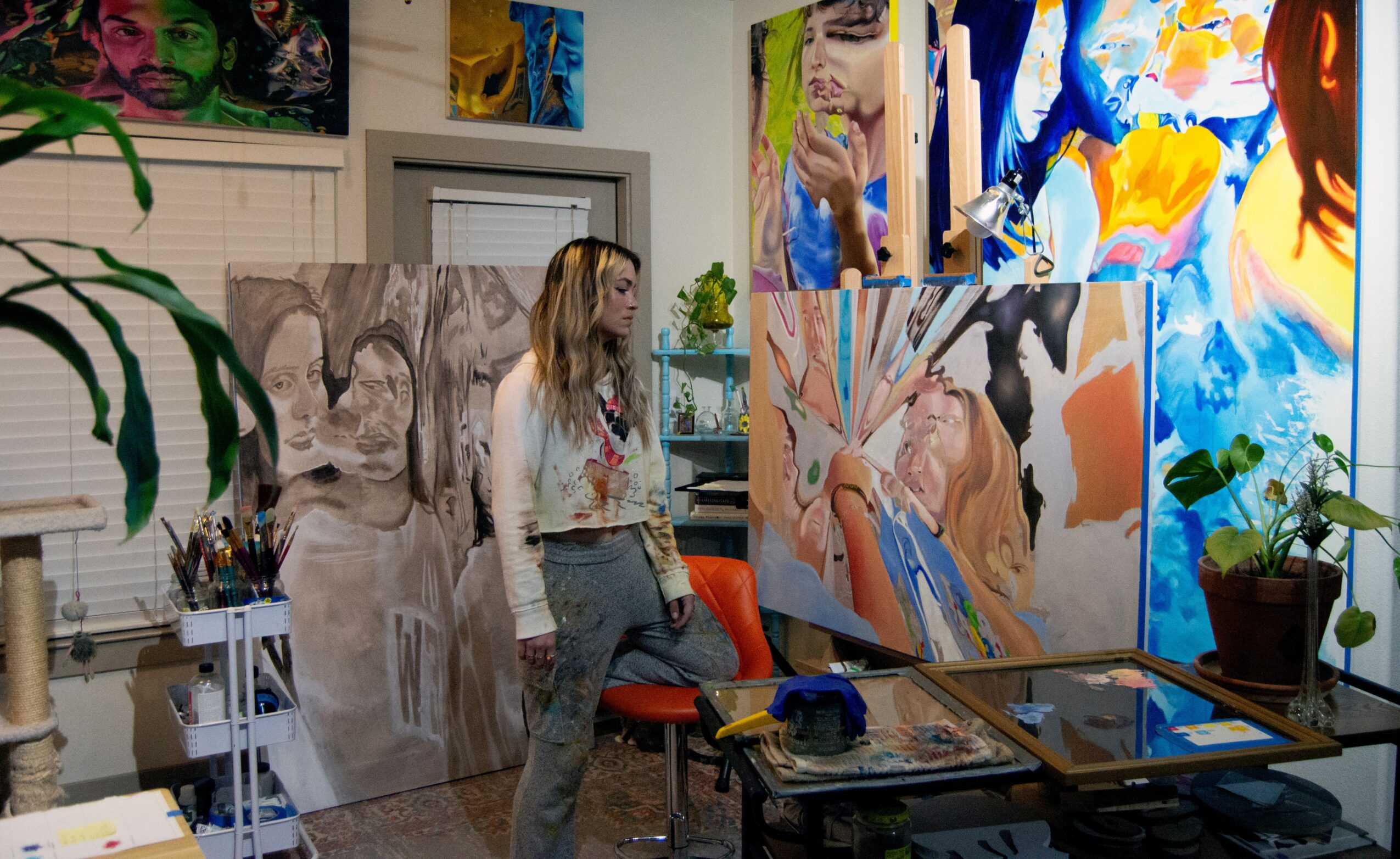 Woman in art studio surrounded by colorful paintings and art supplies, with plants and furniture nearby.