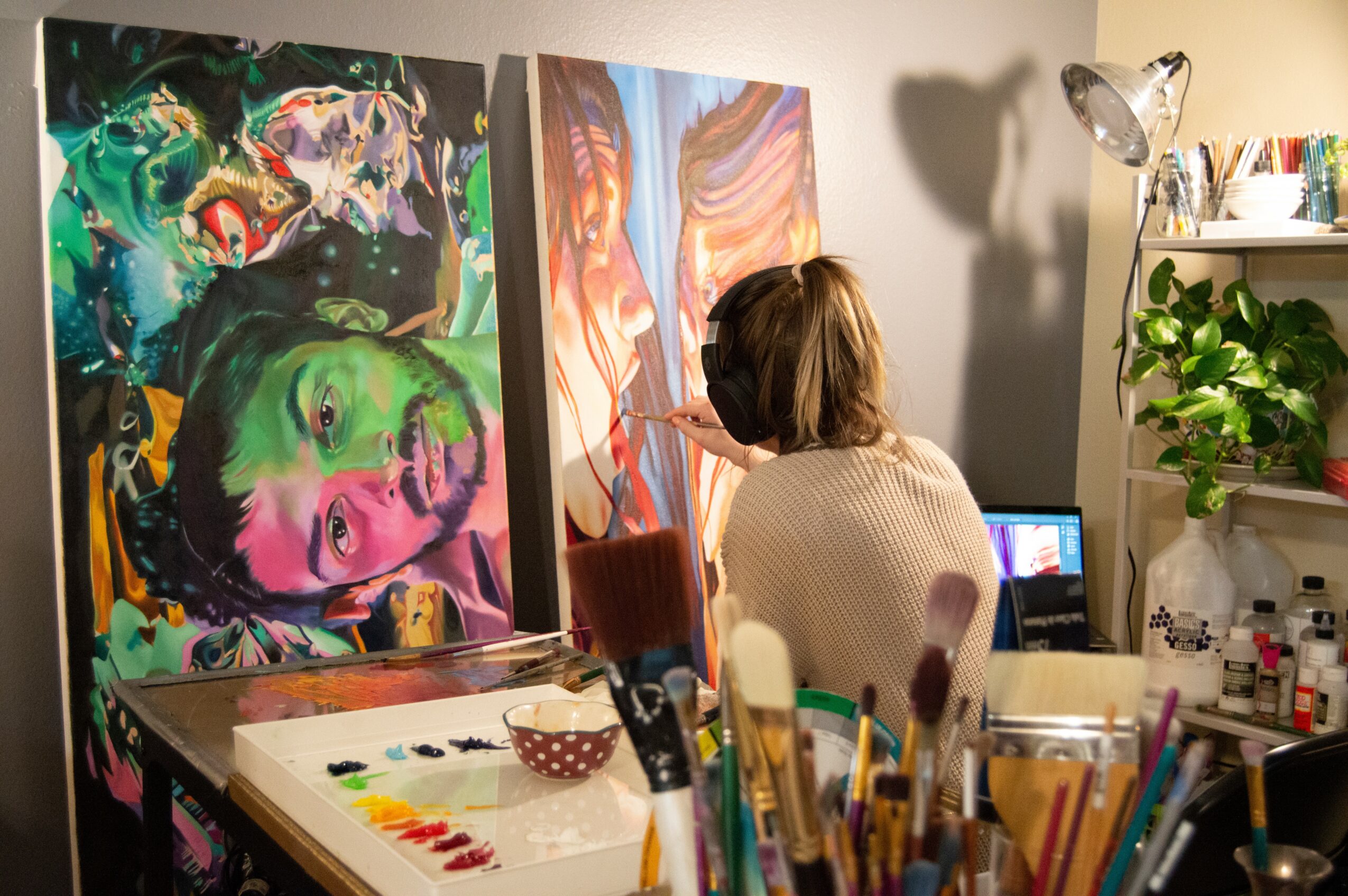 Person with short hair painting in an art studio with colorful paintings, brushes, and a laptop nearby.