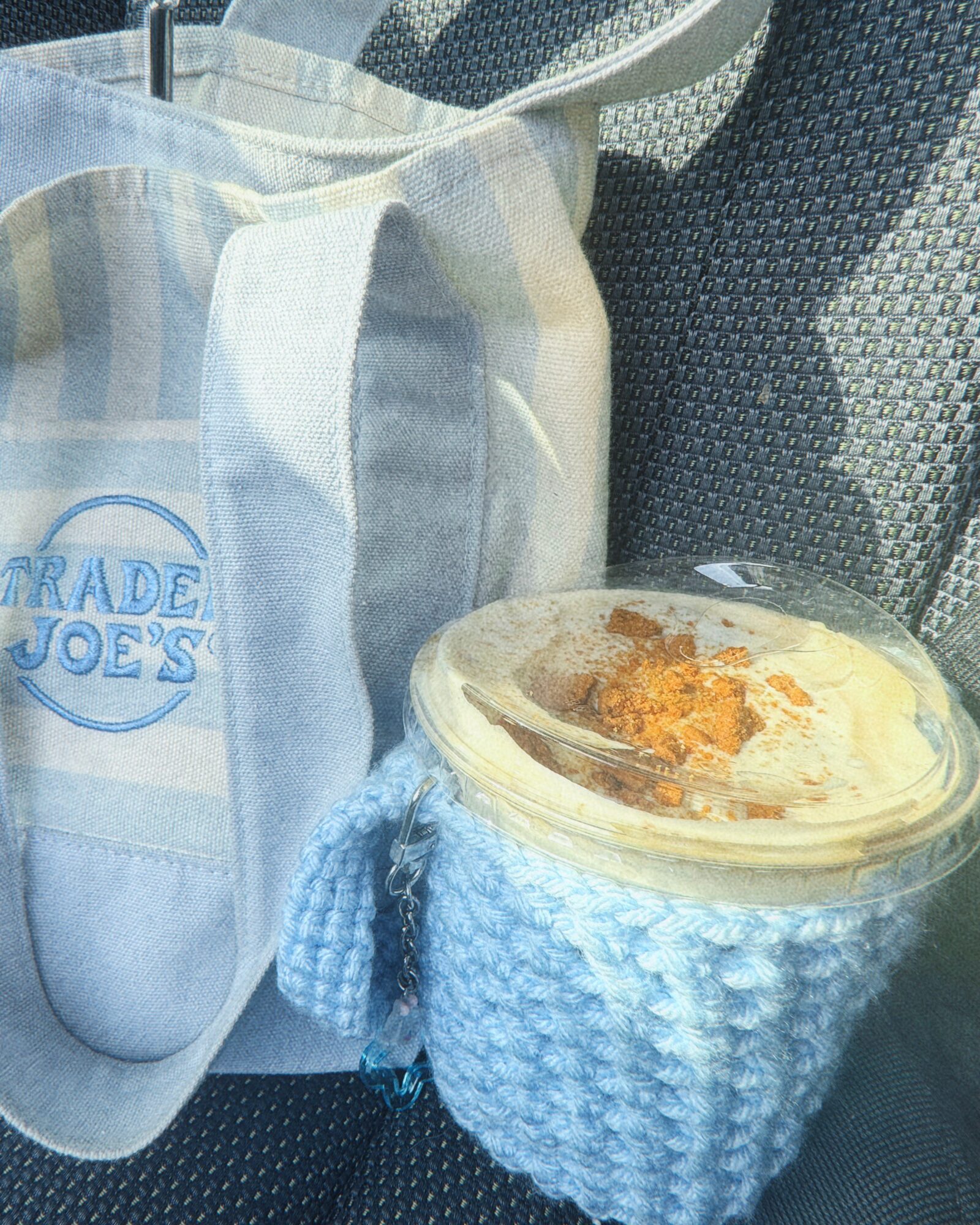 A cup of iced coffee with whipped cream and caramel drizzle in a knitted cozy, next to a tote bag on a car seat.