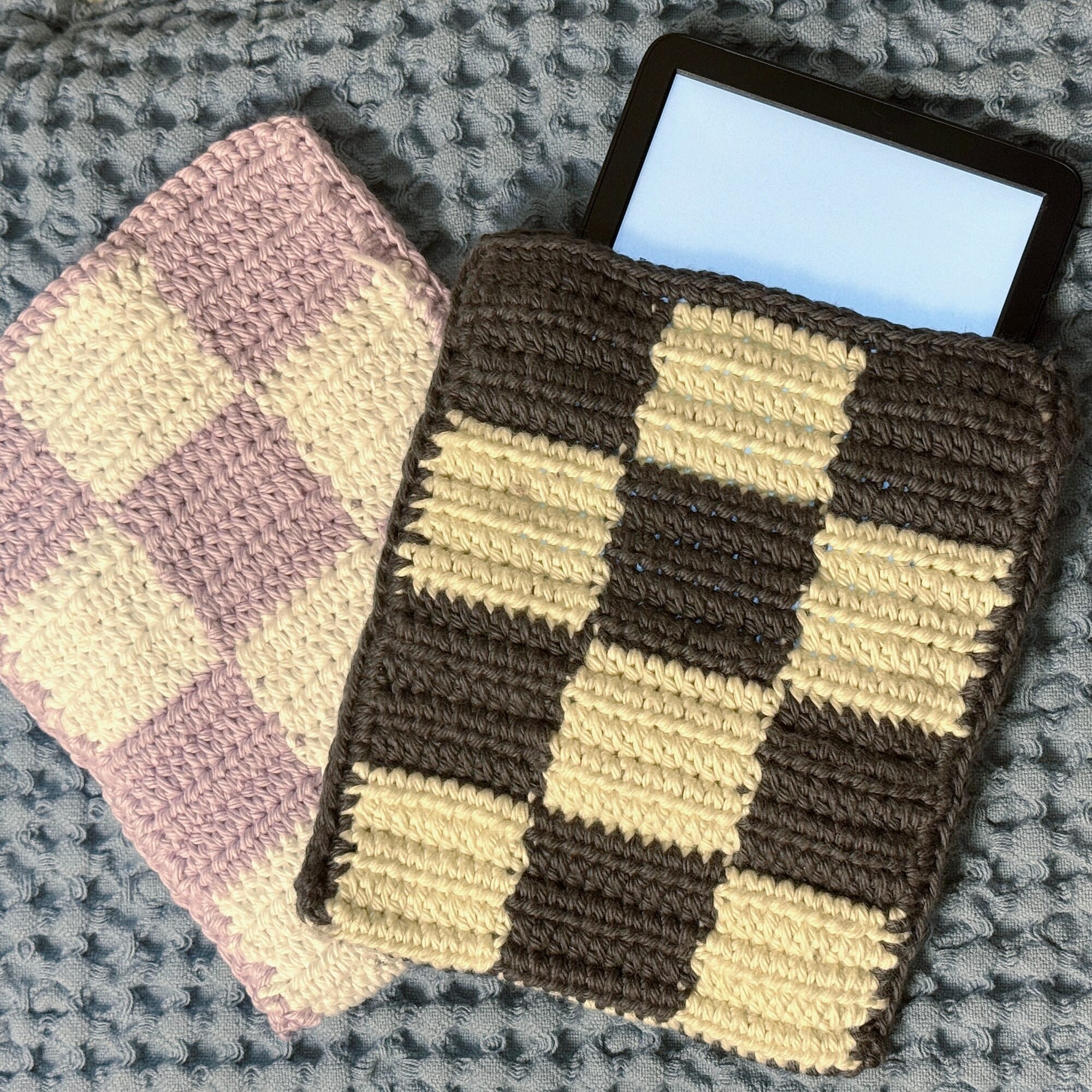 Two knitted tablet sleeves with checkered patterns, one pink and cream, the other brown and cream, on a textured gray surface.
