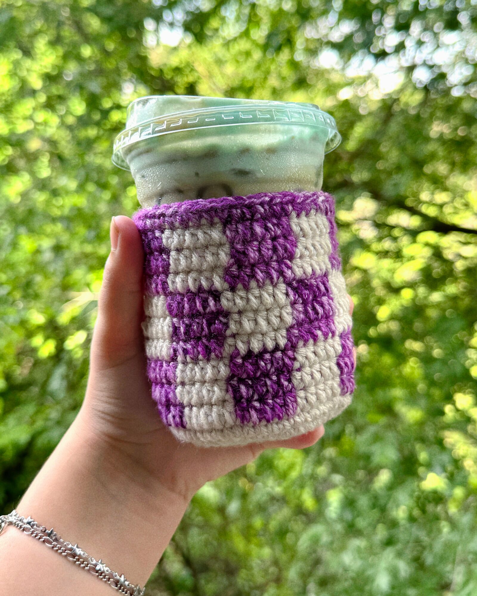 Hand holding a cup with a purple and white crocheted cozy, outdoors with green trees in background.