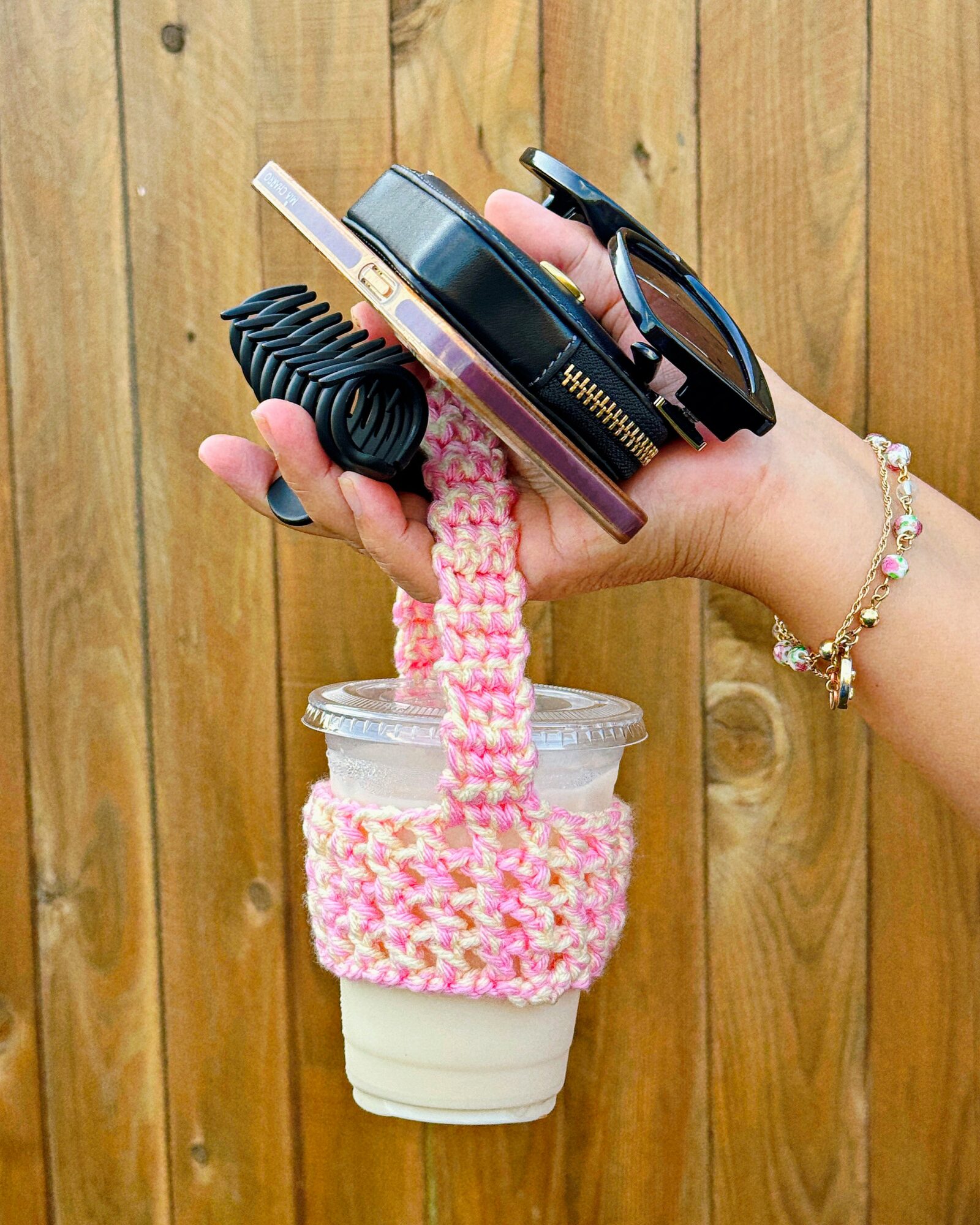 Hand holding a camera, sunglasses, and a pink crocheted cup holder with a drink, against a wooden background.