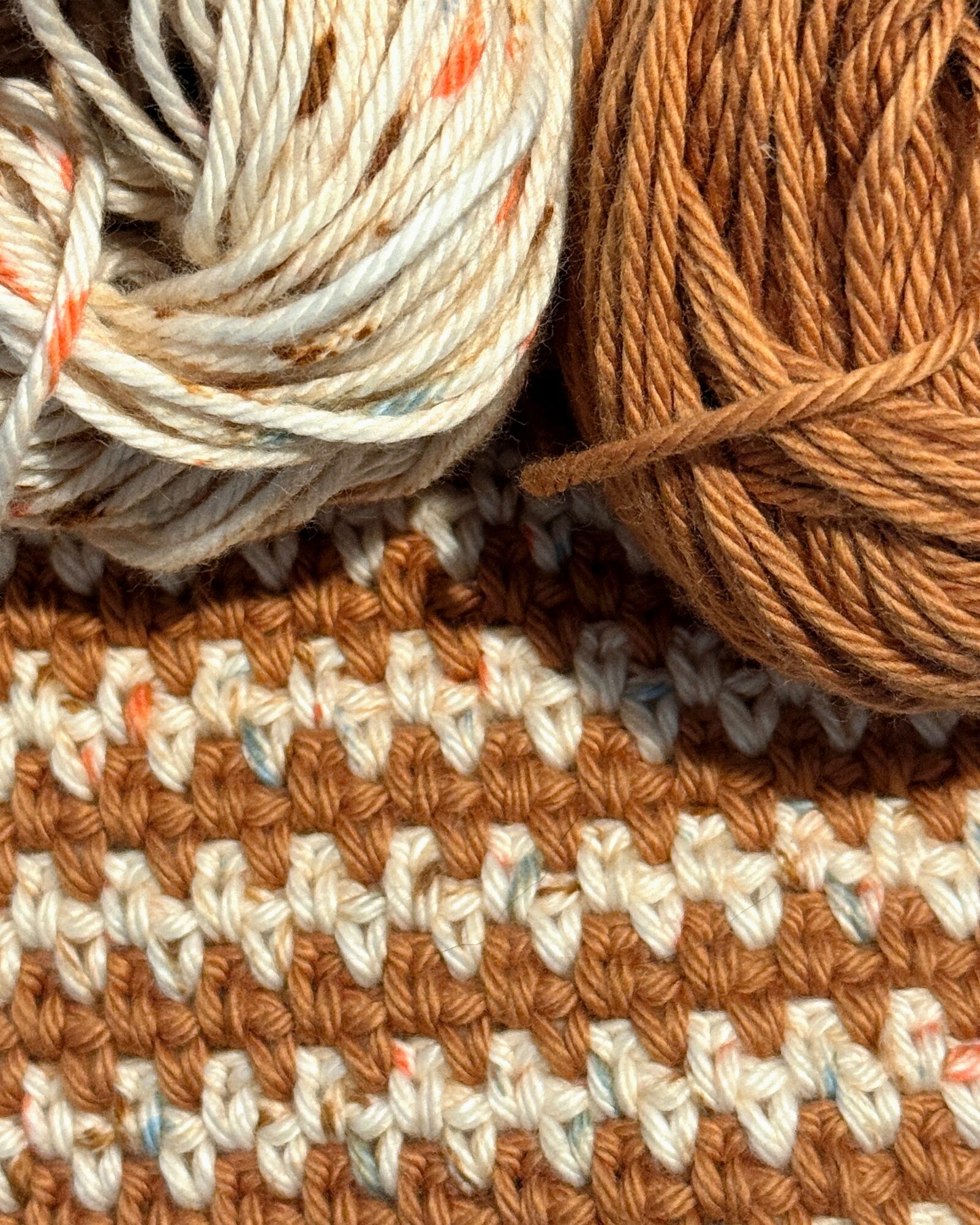 Close-up of two balls of yarn, one cream with orange specks and one solid brown, on a knitted fabric background.
