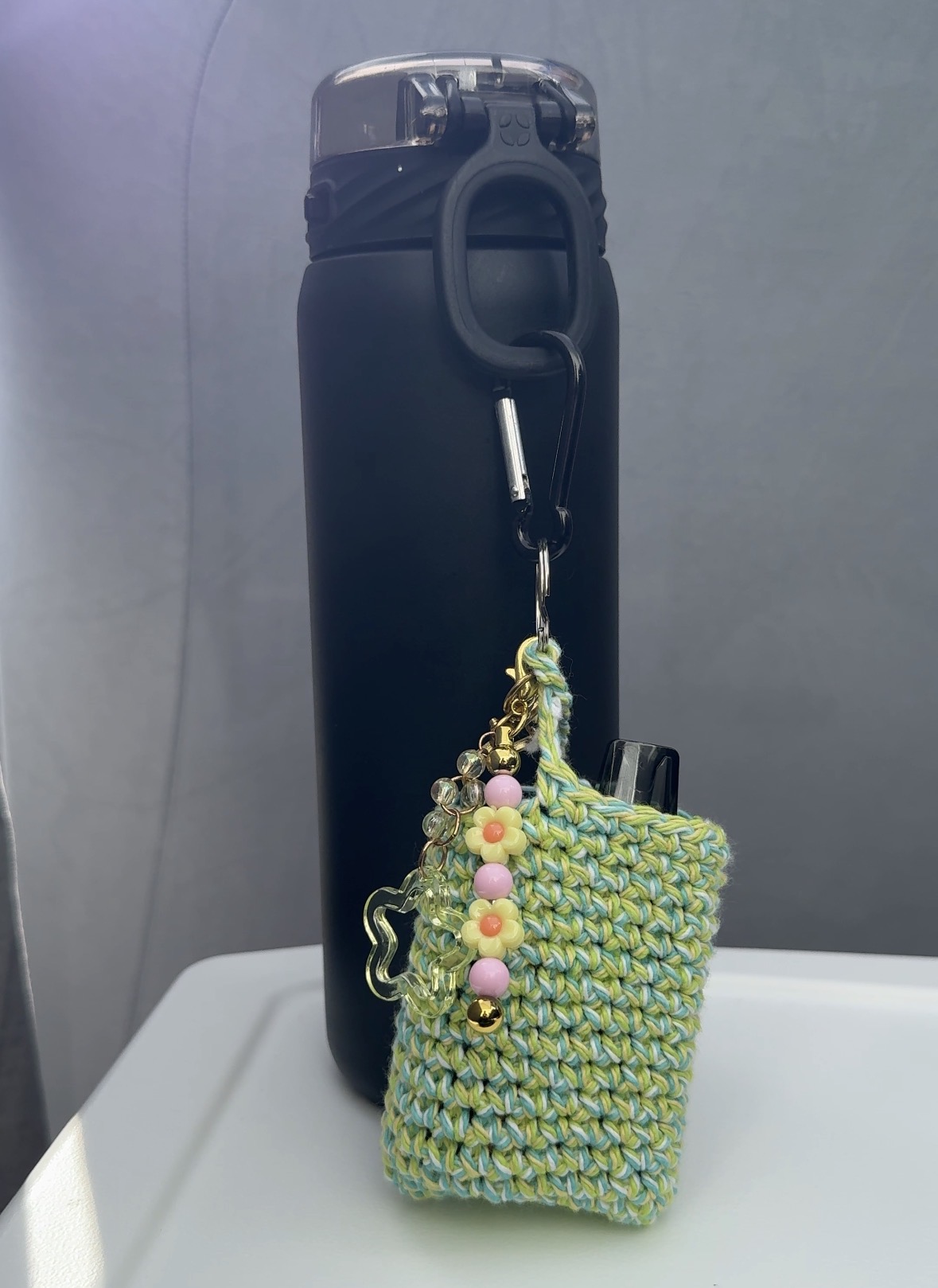 Black water bottle with a small green woven pouch hanging from its handle, on a white surface against a gray background.