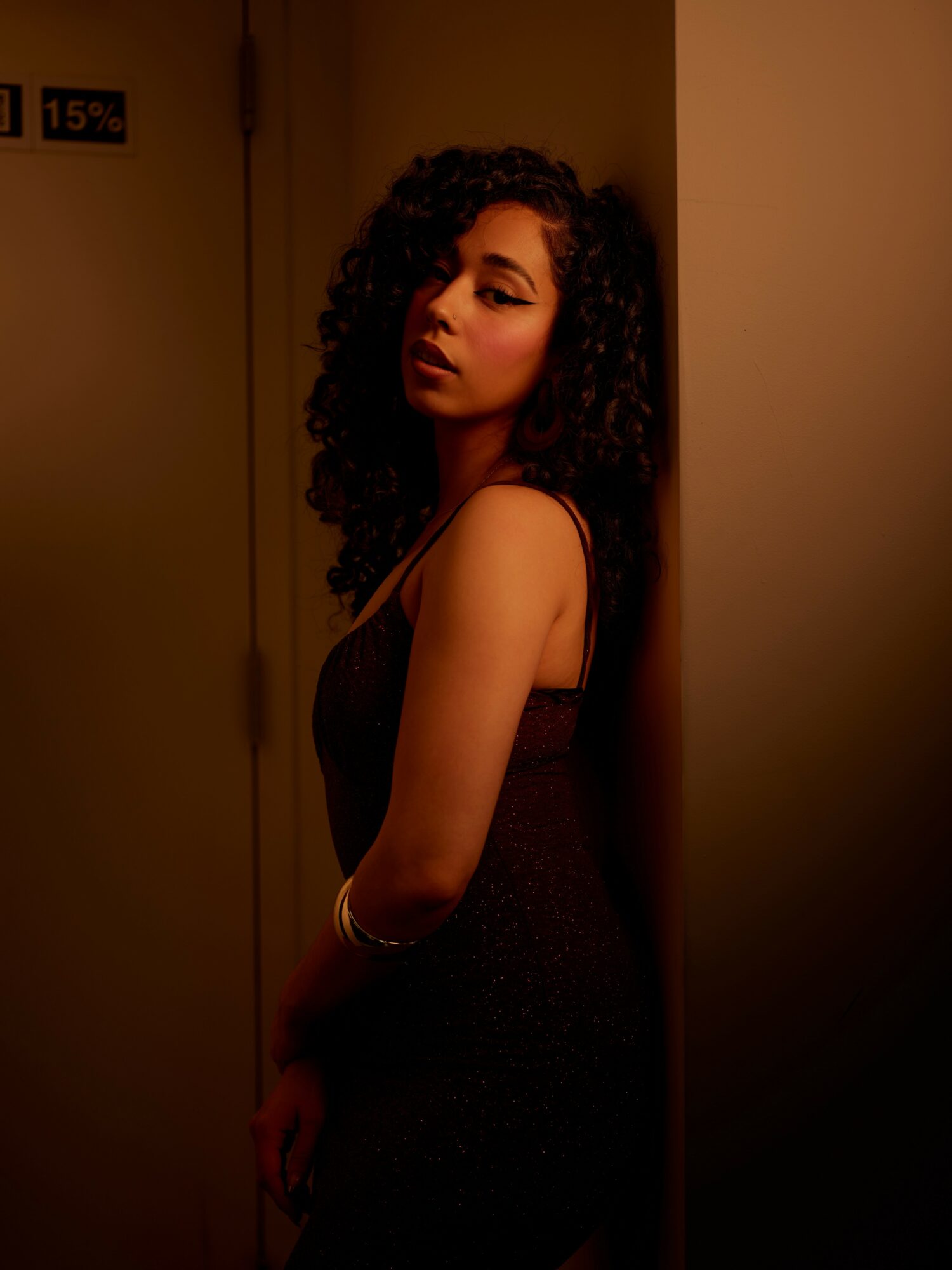 Young woman with curly hair leaning against a wall in dim lighting, looking at the camera, wearing a sleeveless dress.