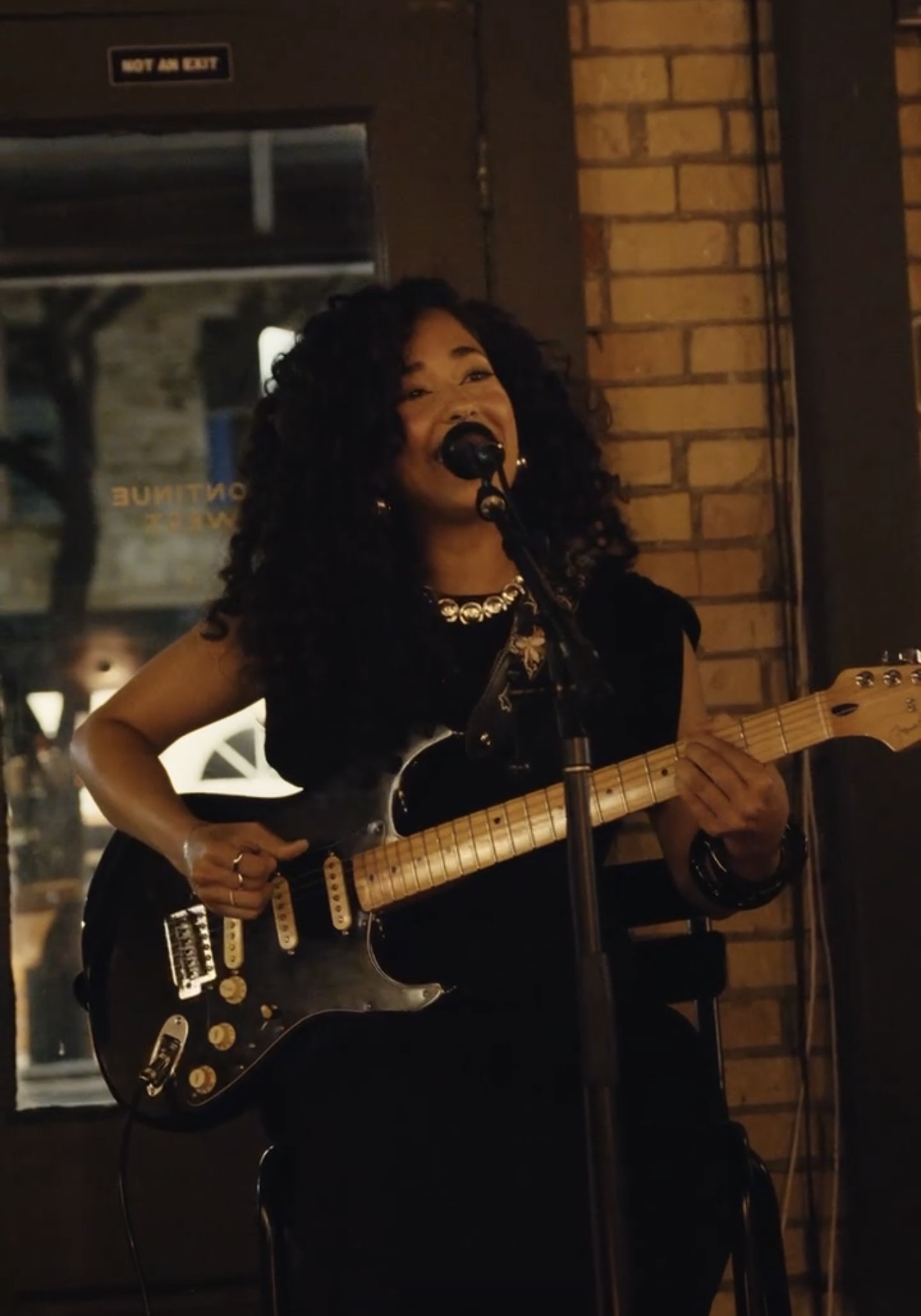Woman with curly hair singing into microphone while playing electric guitar, standing indoors near brick wall.