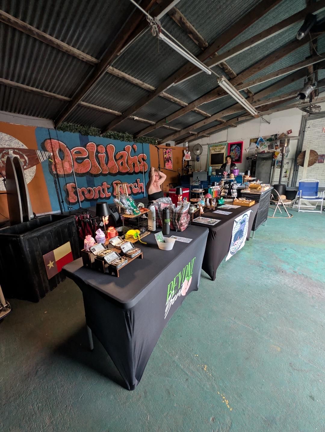 Indoor event space with tables displaying various items, a large colorful sign, and a corrugated metal ceiling.