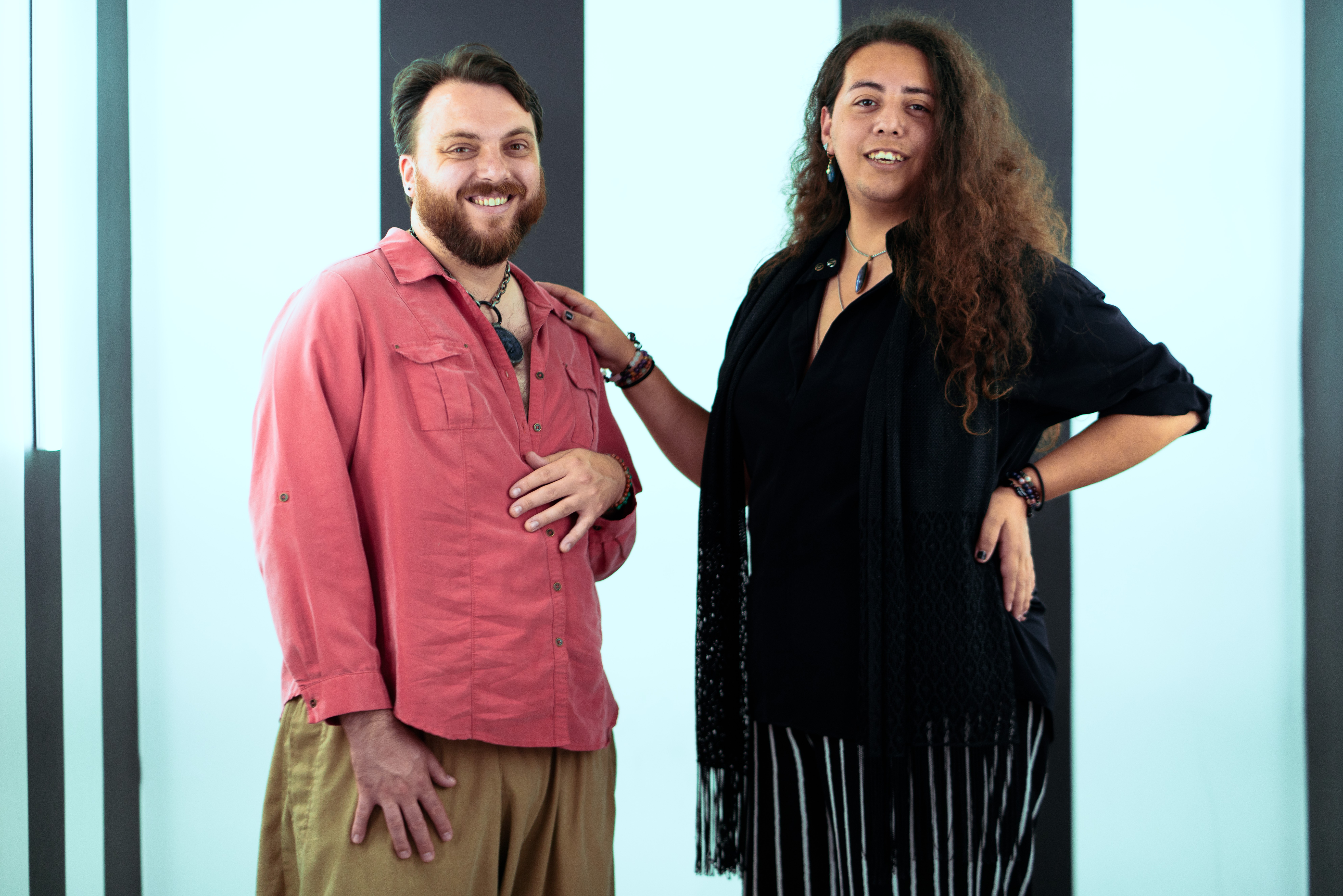 Two people standing together, smiling, in front of a light blue background, one with a beard and the other with long curly hair.