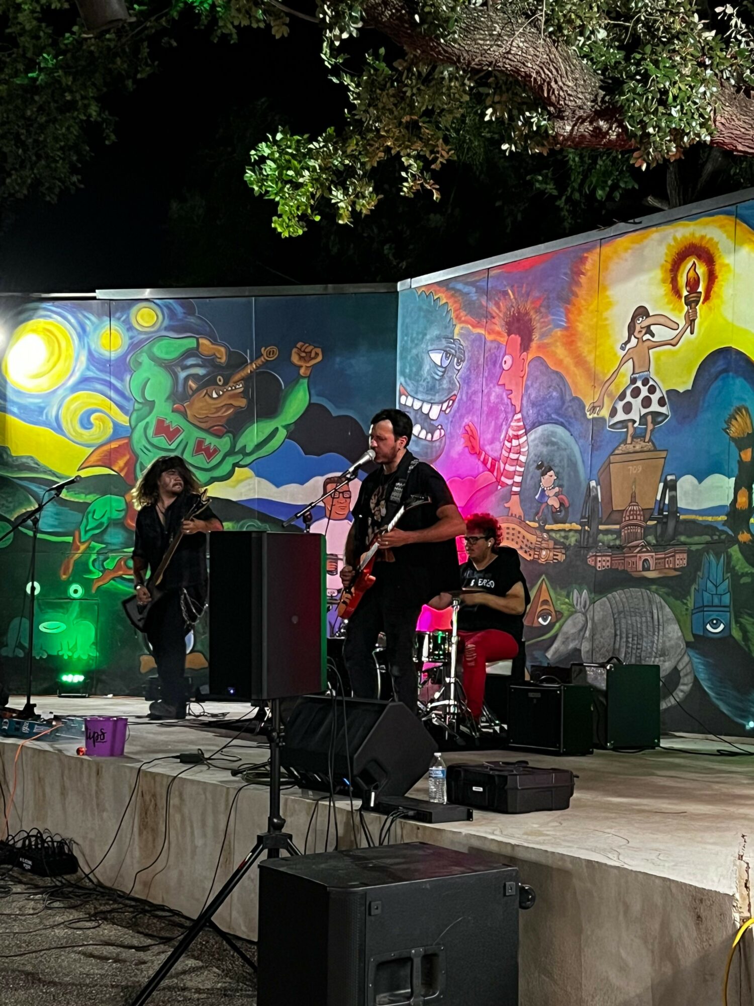 Musicians perform on stage with colorful mural background, playing instruments and singing, with sound equipment and stage lighting visible.