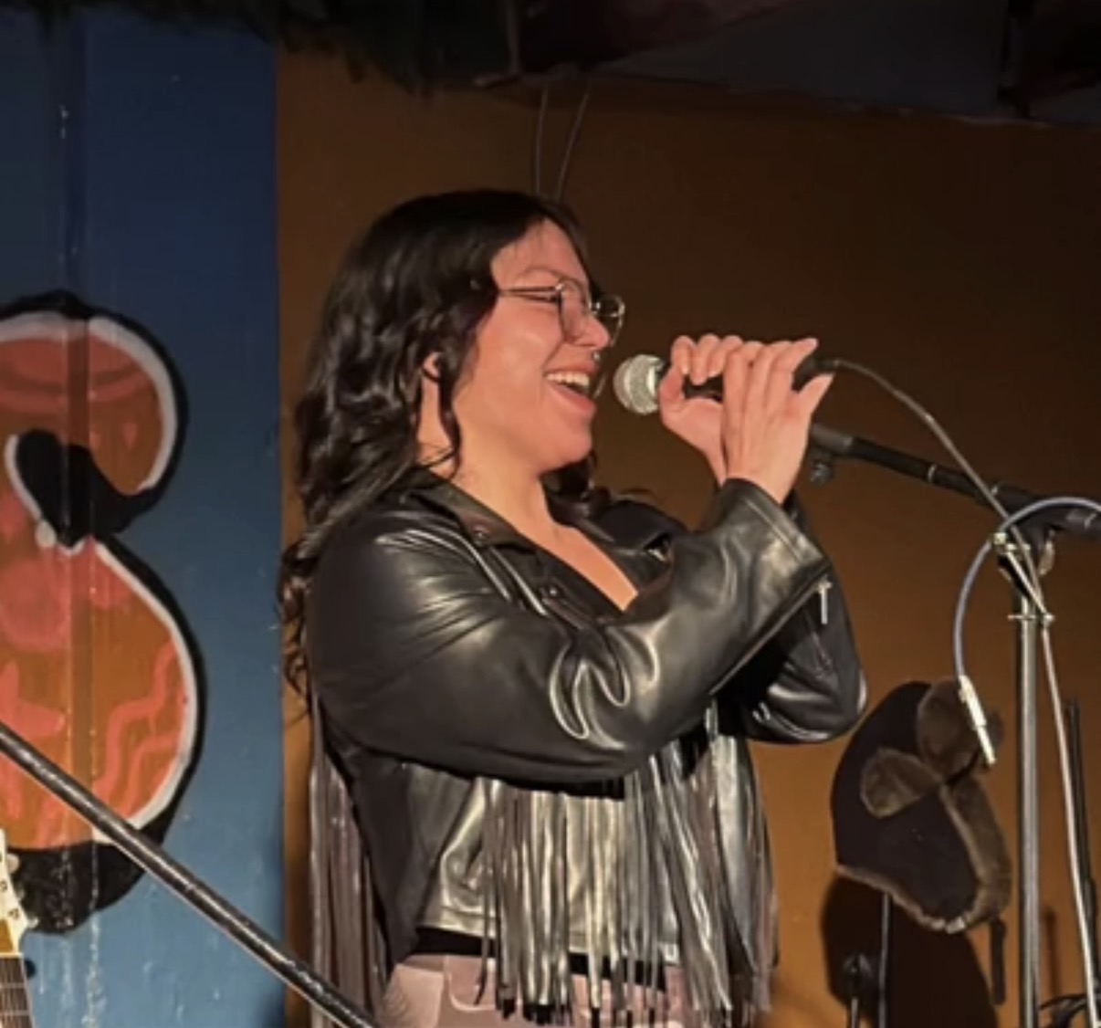 Woman singing into a microphone on stage, wearing glasses and a leather jacket, smiling, with colorful background.