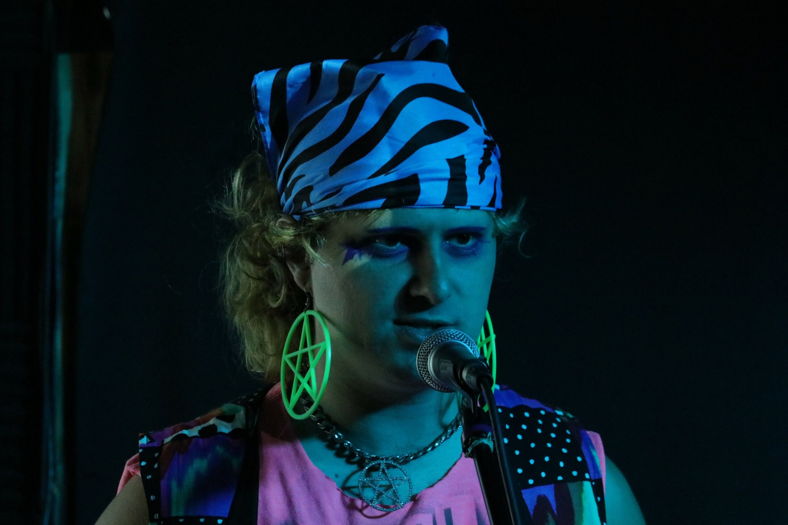 Person with zebra-striped headscarf, large green earrings, and colorful clothing speaking into a microphone against a dark background.
