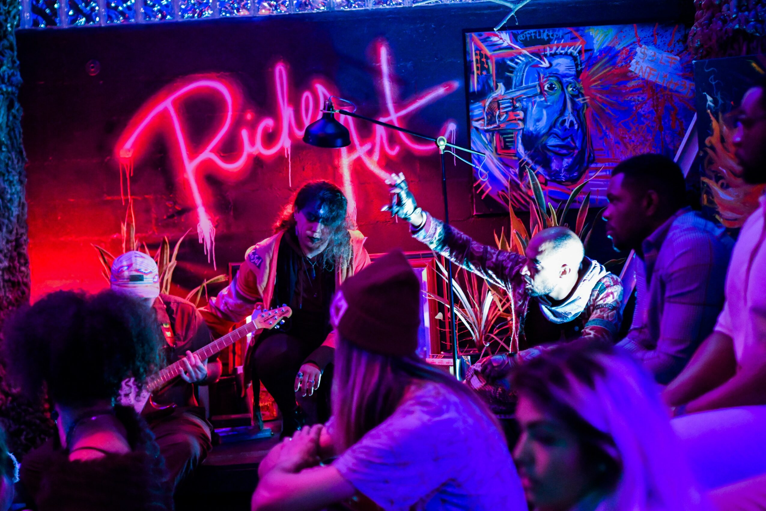 Group of people sitting and playing guitars in a neon-lit room with graffiti art and a pink neon sign that says