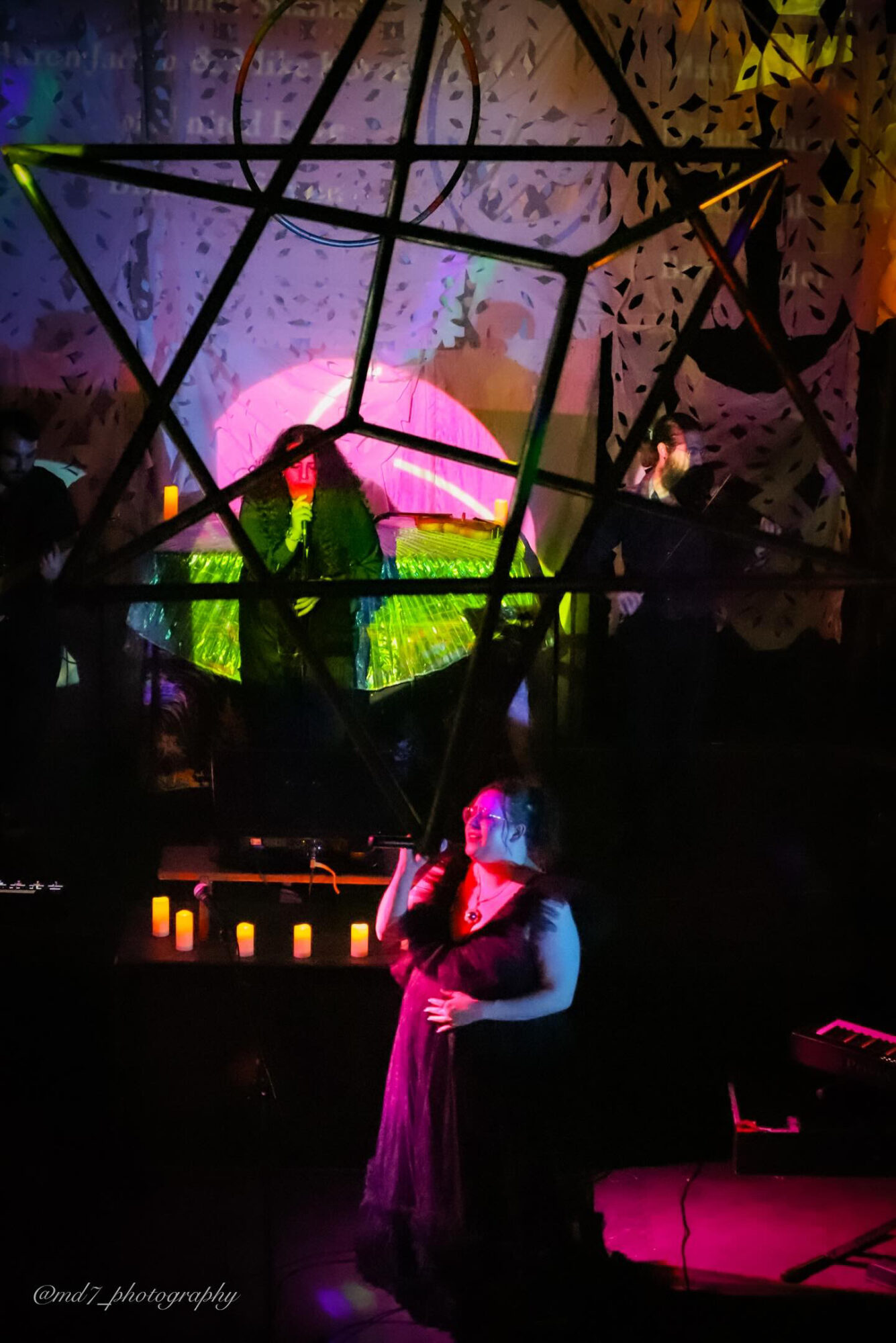 Performer singing on stage with colorful background and silhouettes of audience, illuminated by pink and purple lights.