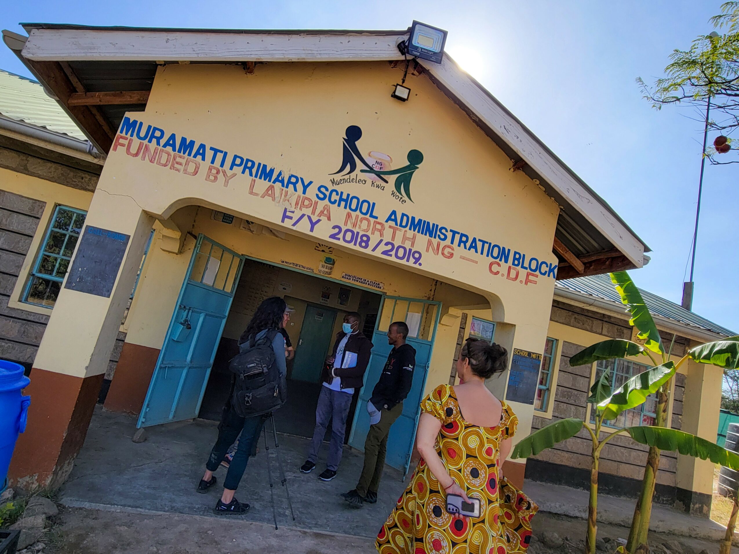 Primary school building with a sign, people standing outside, and a banana plant nearby.