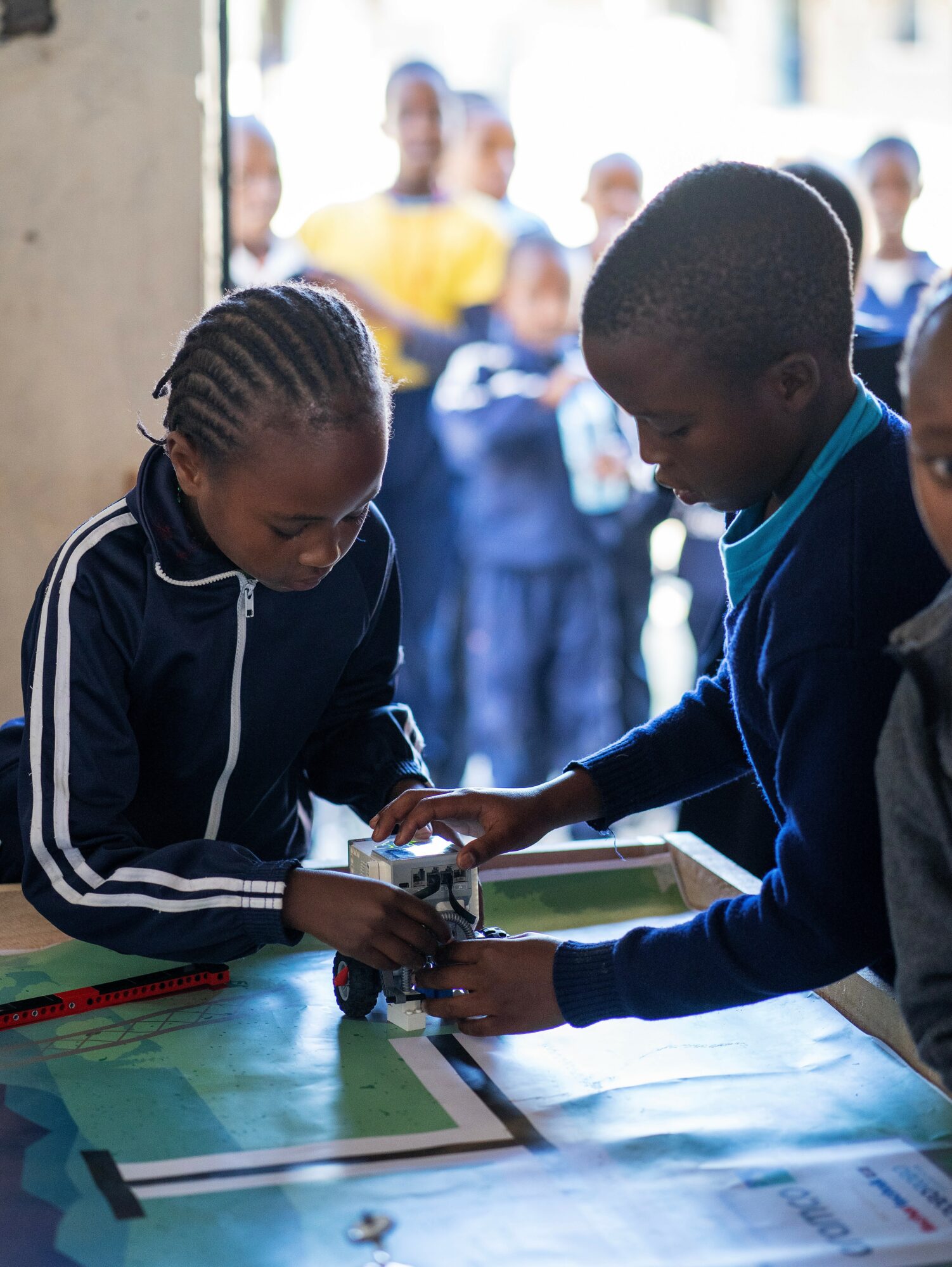 Two children working together on a project at a table outdoors, with a group of people in the background.