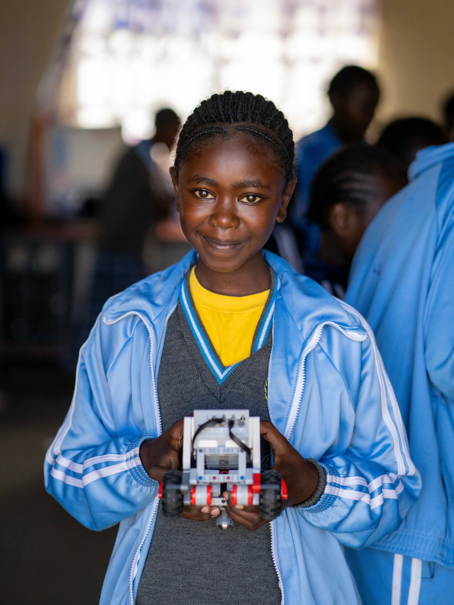 Young girl smiling, holding a small robot, wearing a blue jacket and yellow shirt, in a classroom setting.
