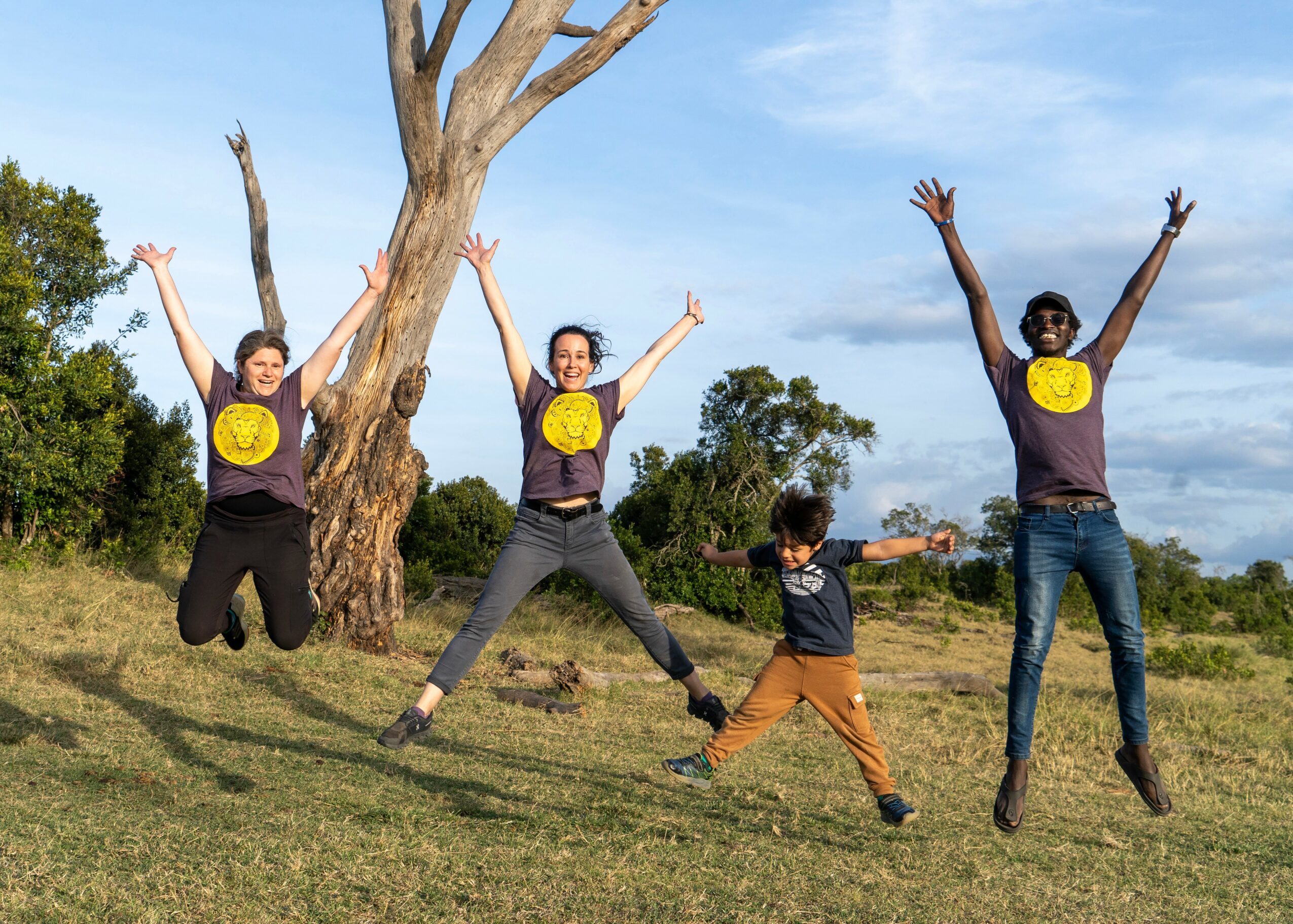Four people, three adults and one child, jumping outdoors with arms raised, under a blue sky with trees and grass.