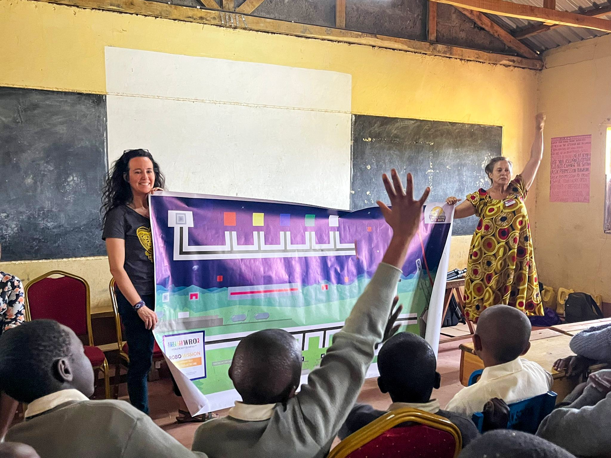 Two women are presenting to a group of children in a classroom, with one woman raising her hand and the other standing near a colorful display board.