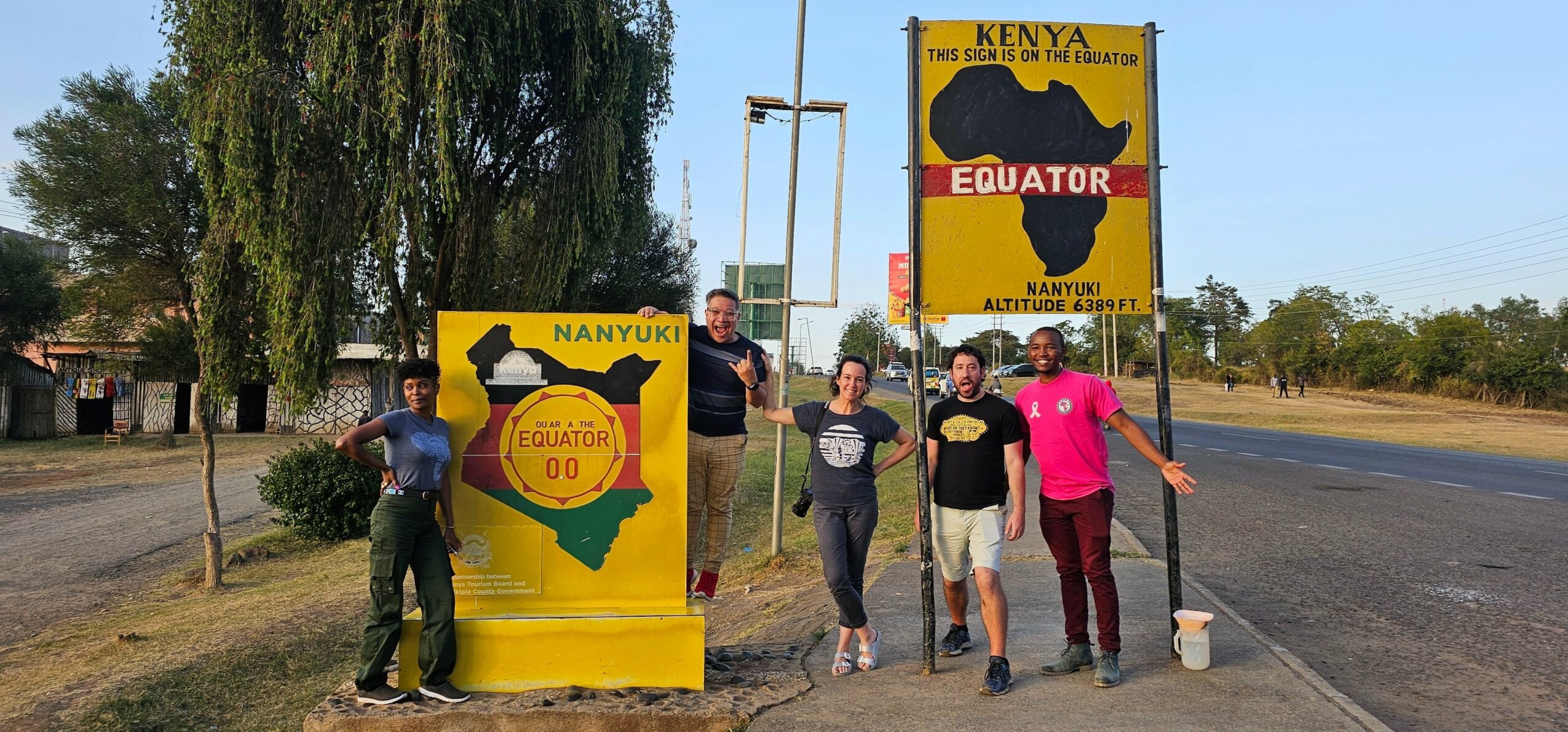 Group of five people standing near signs marking the Equator in Kenya and Nanyuki, with trees and a road in the background.