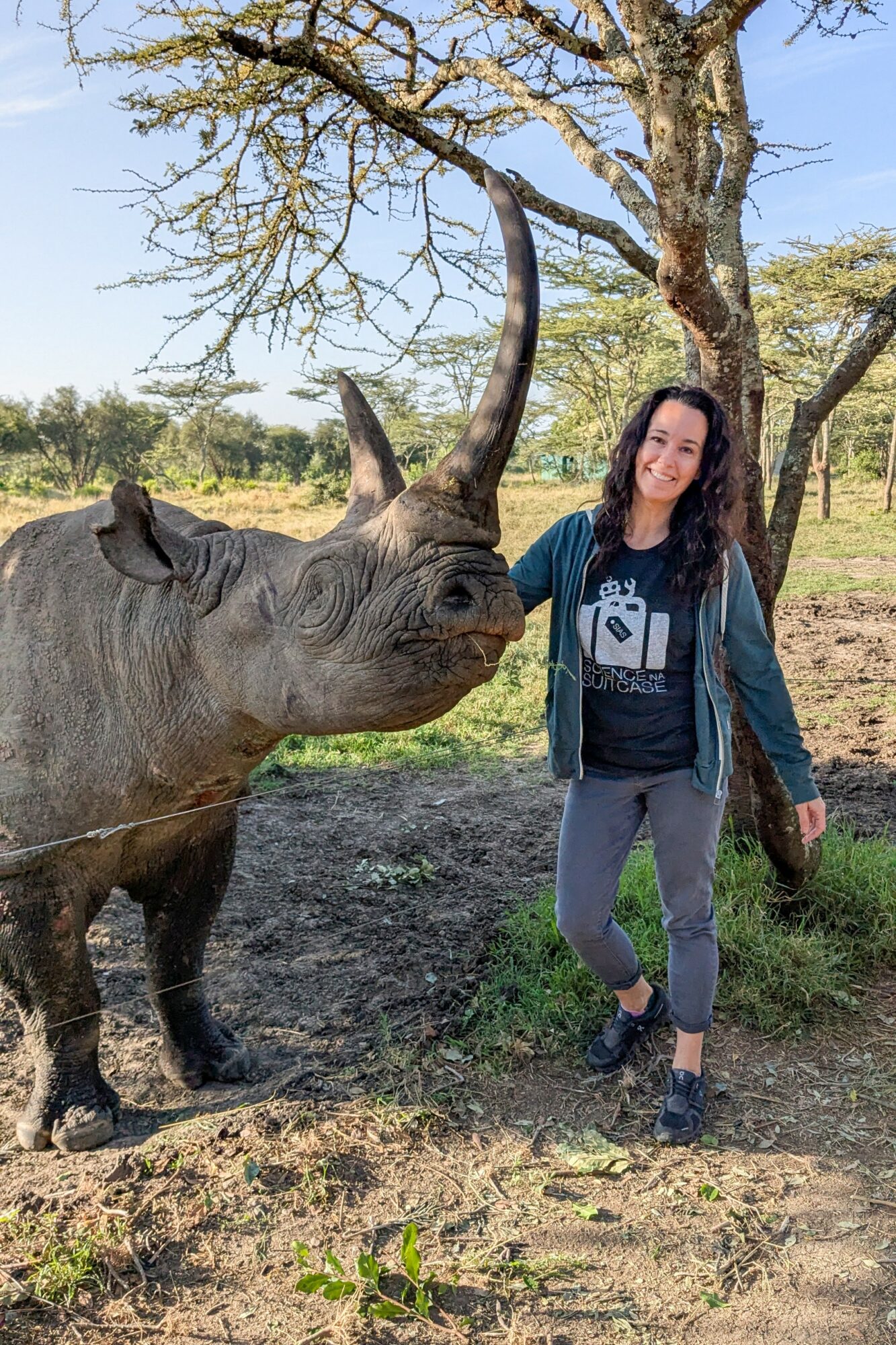 Woman standing next to a rhinoceros outdoors with trees and grass, smiling at the camera.