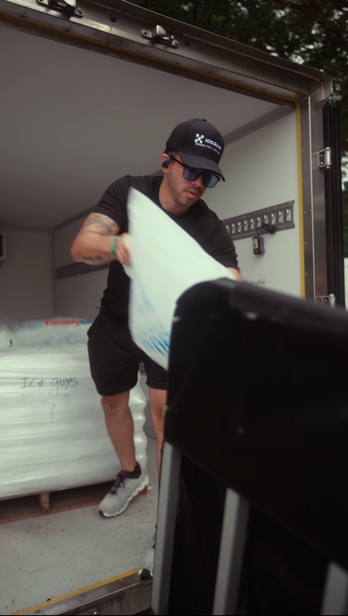 Person wearing a black cap and sunglasses, holding a surfboard inside a truck, with a bed and hooks in the background.