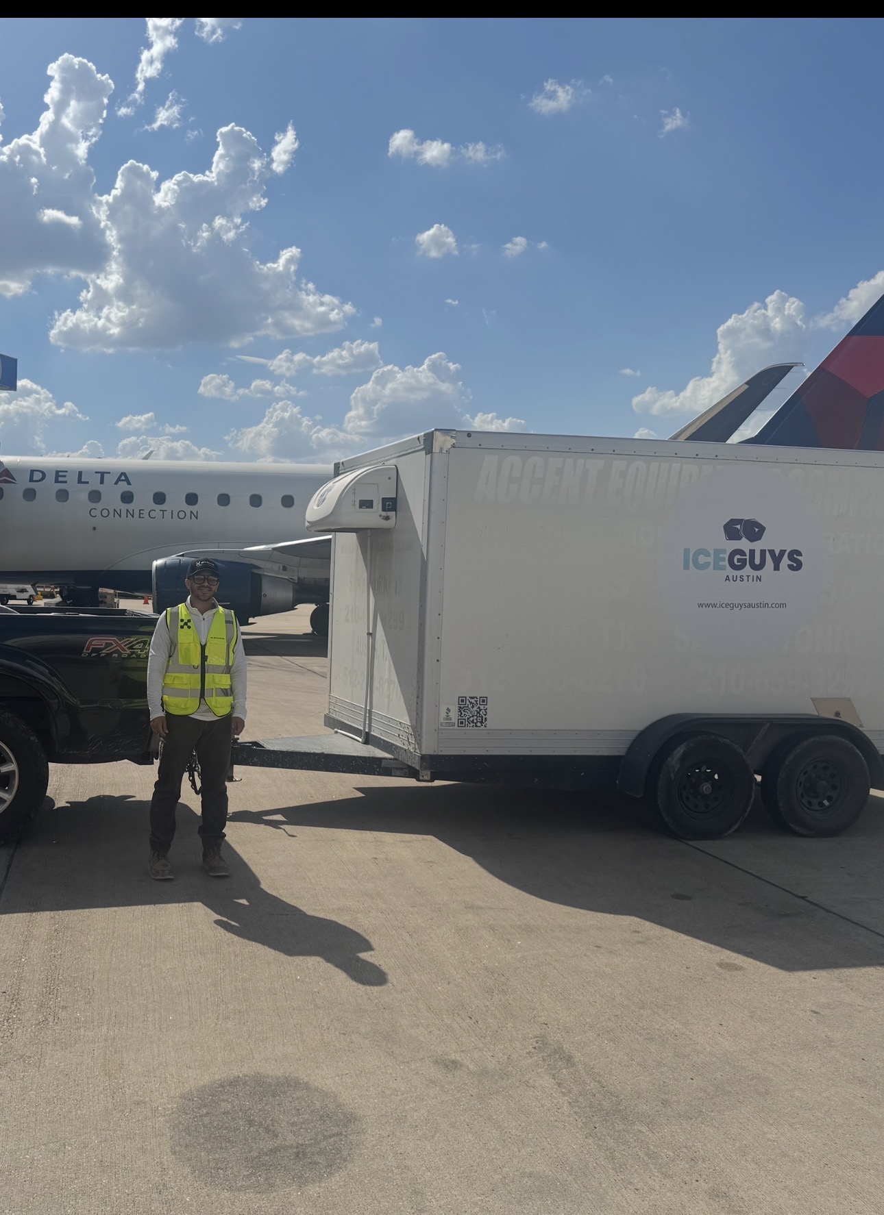 Person in a yellow safety vest standing next to a mobile ice cream cart on airport tarmac, airplane in background, partly cloudy sky.