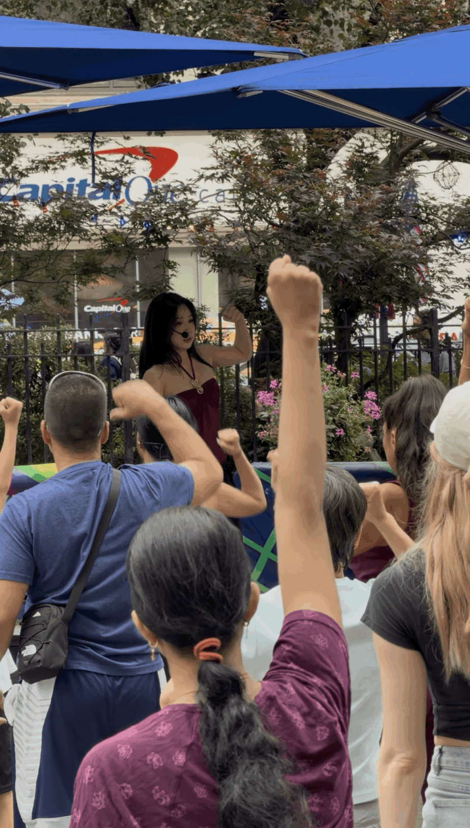 Woman speaking to crowd outdoors with raised hand, people listening, trees, and a building in background.