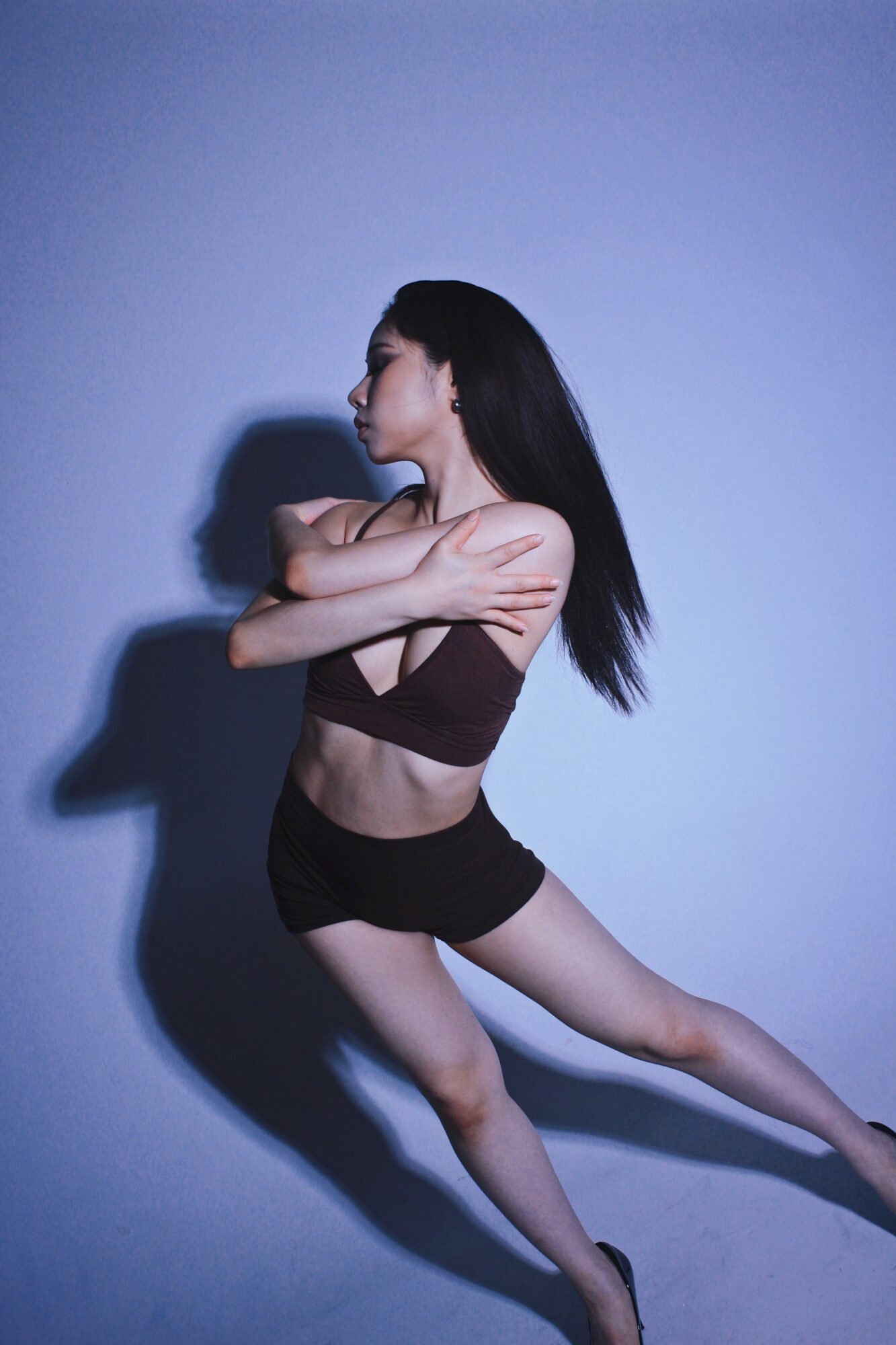 Woman in black sportswear stretching with arms crossed, shadow cast on wall, side profile, dark hair, plain background.