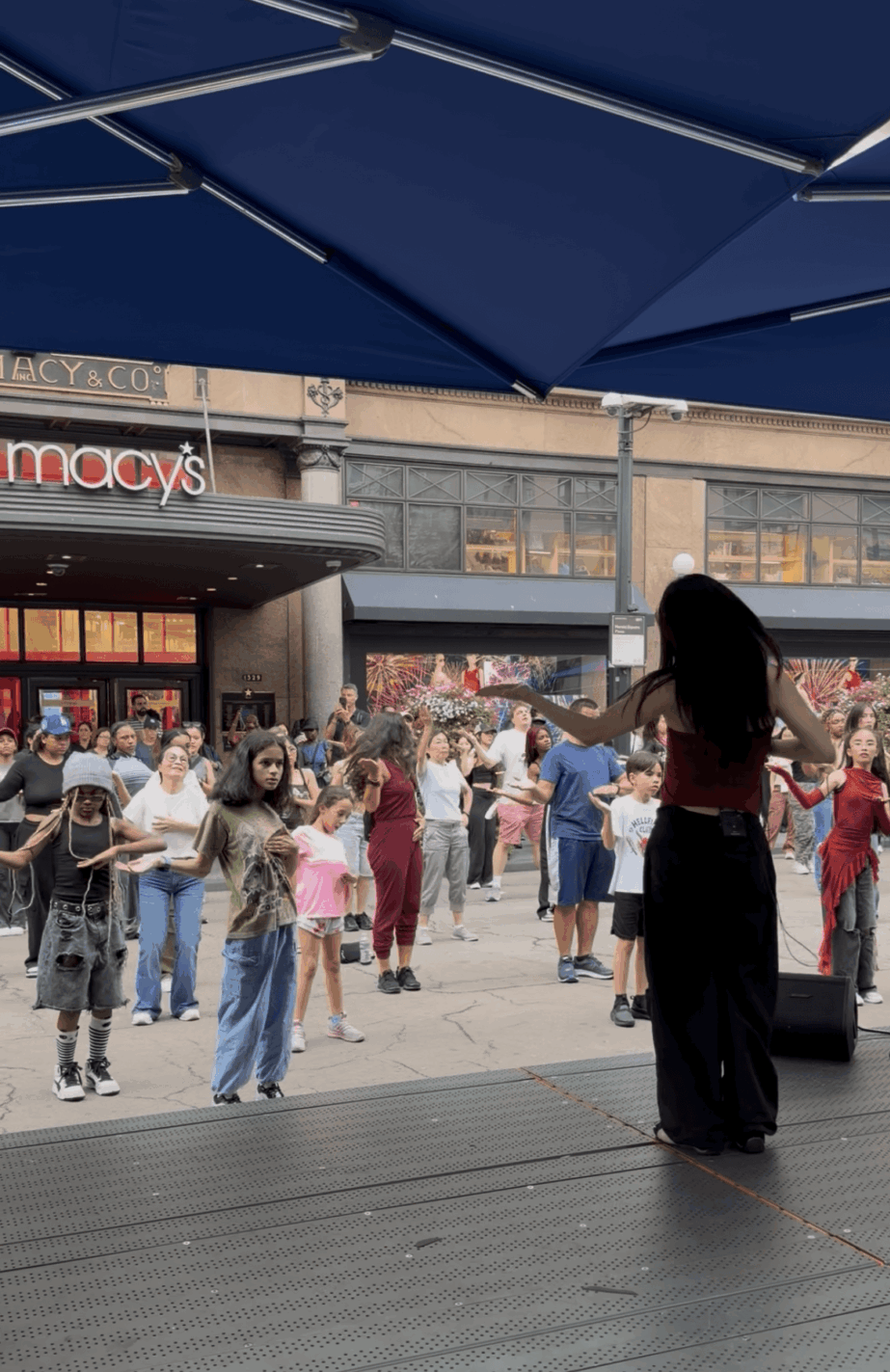 People gather outside a building with a store and large windows, under a blue canopy, with a person standing in the foreground.