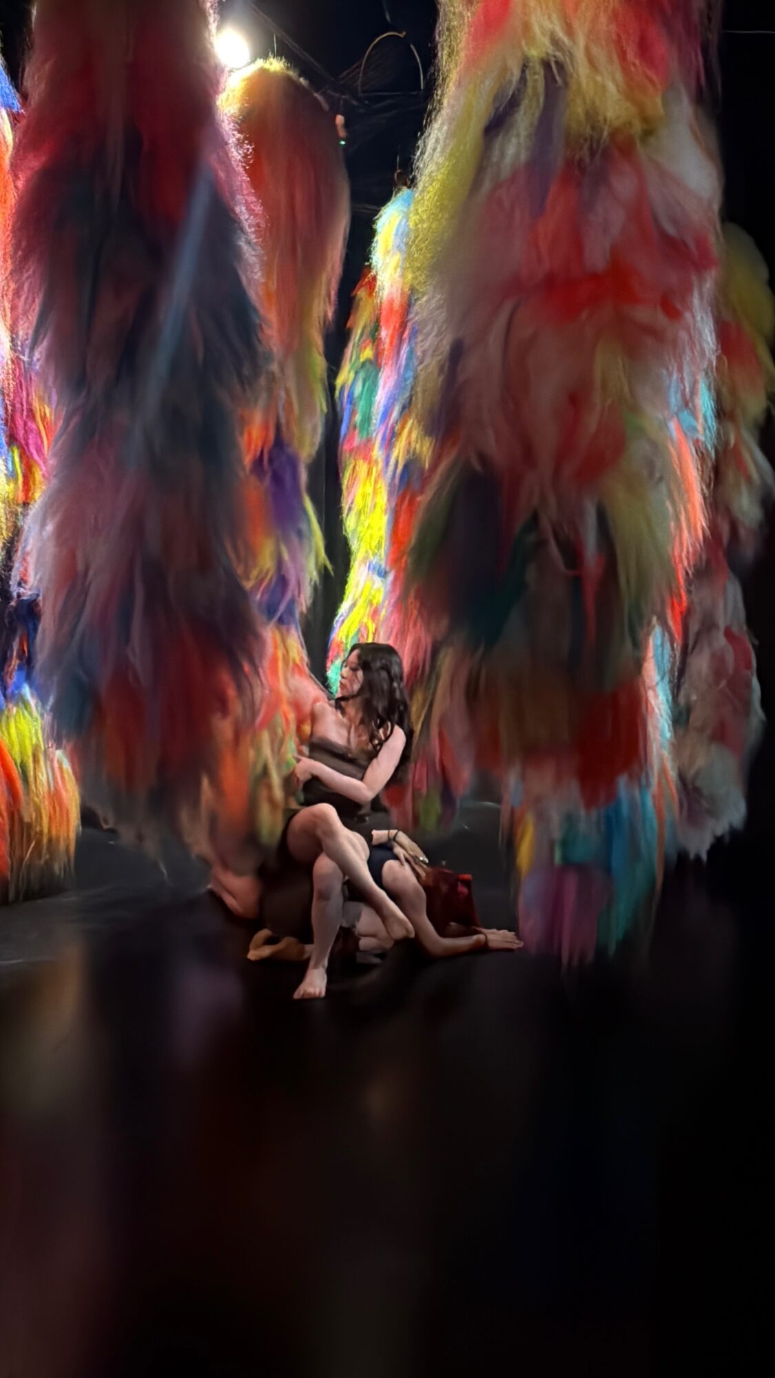 Person sitting on the floor surrounded by large, colorful, feather-like structures in a dark environment.