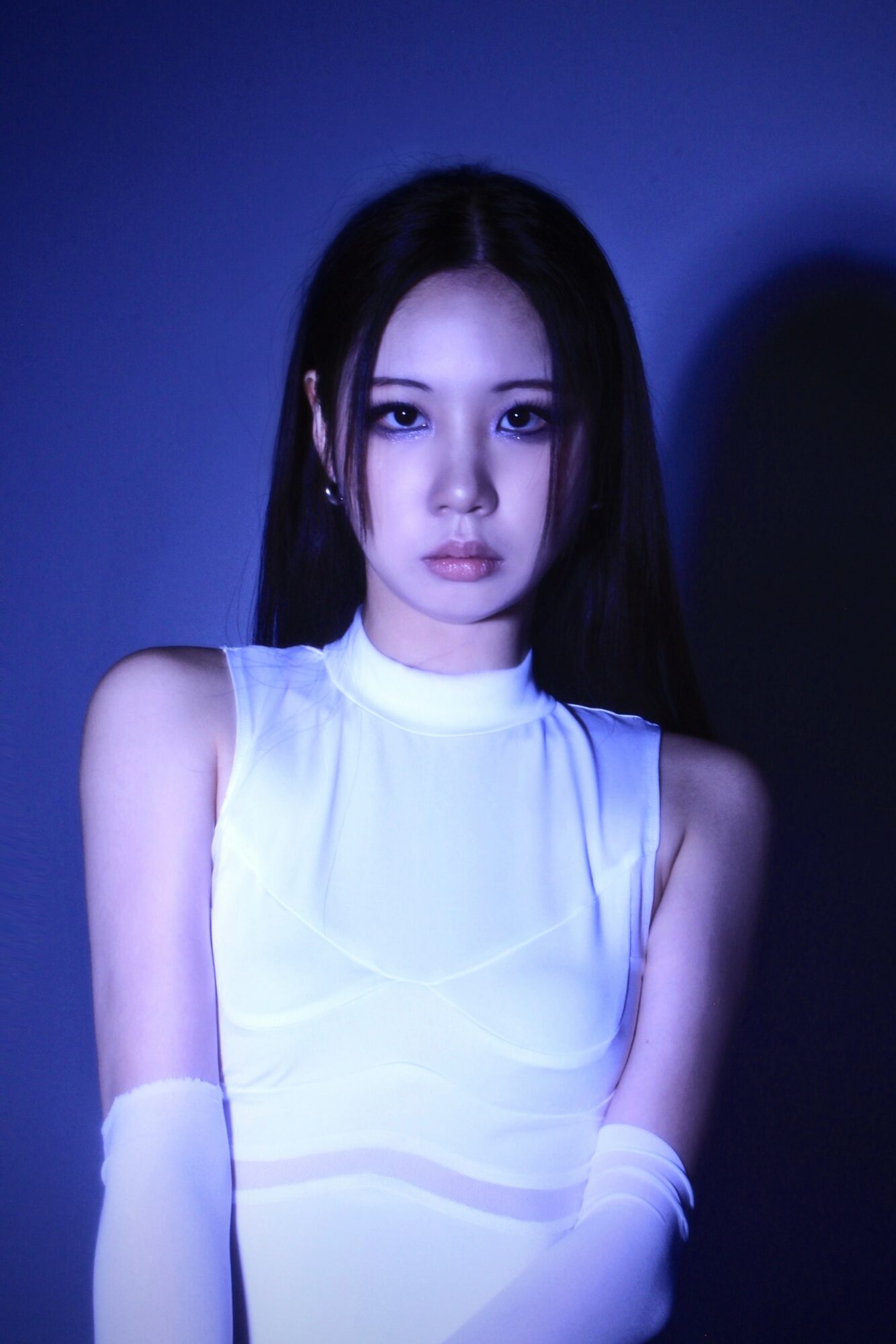 Young woman with long dark hair wearing a white sleeveless top and gloves, looking at the camera against a dark background.