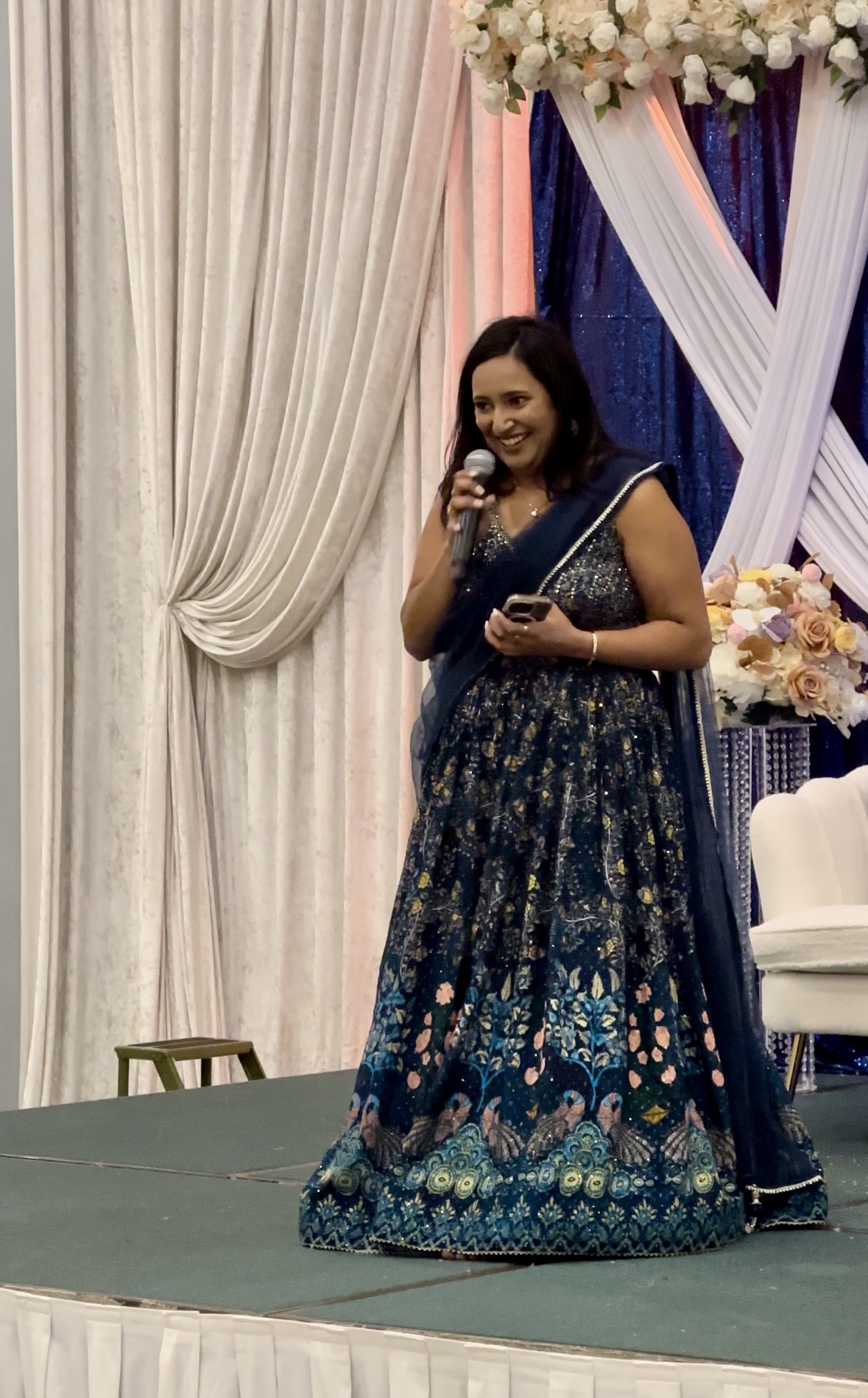 Woman in a blue and gold dress holding a microphone on stage with floral decorations and drapes behind her.