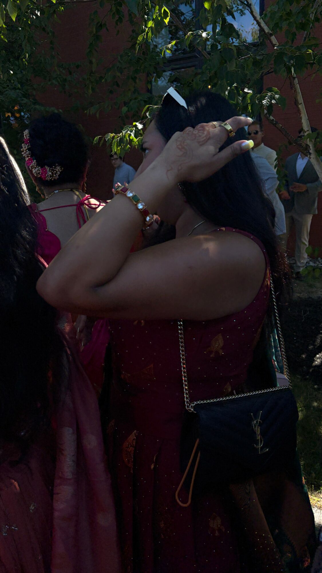 Person adjusting hair outdoors, wearing a maroon dress and carrying a black purse, surrounded by trees and people.